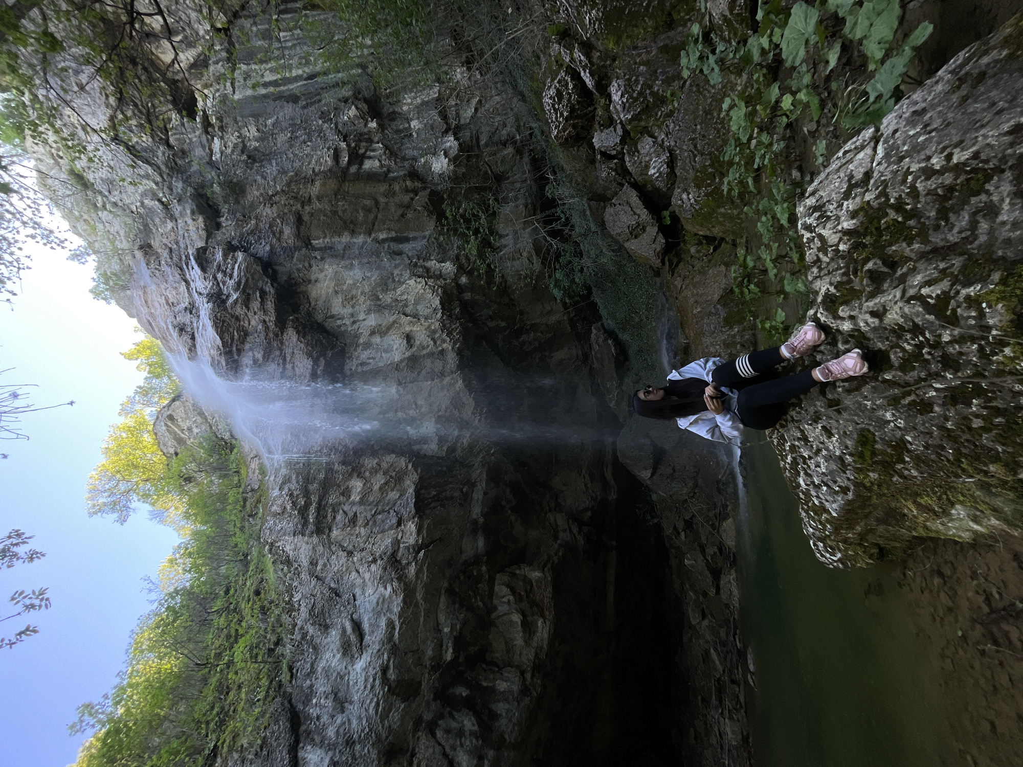 A medium shot shows a person sitting on a moss-covered rock outcrop in front of a waterfall. The person wears a light-colored hoodie with black stripes on the sleeves, black leggings, and pink shoes. They face right, looking toward the waterfall. The waterfall cascades down a steep, rocky cliff face covered with some vegetation. Trees with green leaves are visible at the top of the cliff and on the left side. The waterfall empties into a pool of water. The sky is blue at the top left of the frame. The location is Veselinovo, Bulgaria.