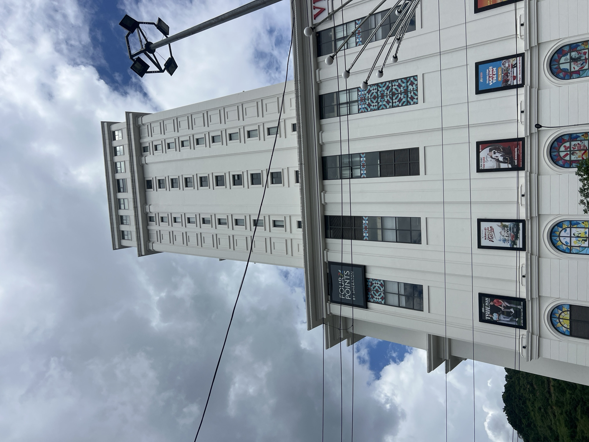 The image shows a tall, white, modern building in Hà Giang 2 Ward, Vietnam, under a cloudy sky. The building has many windows arranged in a grid pattern. A prominent sign for "Four Points by Sheraton" is visible on the building. Several framed advertisements or posters are also displayed on the lower facade of the building. A street light fixture with multiple lamps is positioned to the right of the building. A lush green hillside is visible in the background to the left. Electrical wires crisscross the scene in the foreground.