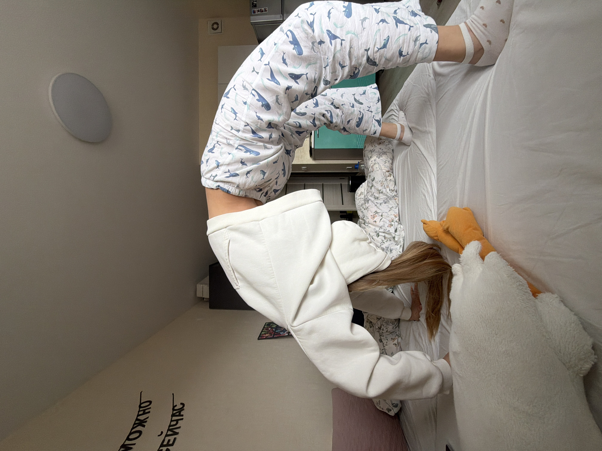 A person in white sweatpants and a white hoodie is bent over backwards in a room, with their hands on a bed. The person is wearing white socks with small hearts on them. The bed has white sheets and a large white and orange plush goose on it. The person also appears to be wearing whale-printed pajama pants. A white circular light fixture is visible on the wall. Text in Russian is visible on the wall: "МОЖНО" and "СЕЙЧАС". The location is potentially Izhevsk, Russia.