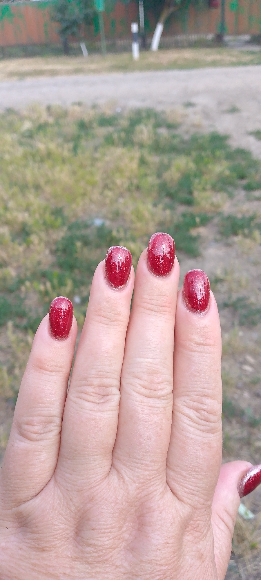 A close-up, top-down view of a left hand shows four nails painted a sparkling, deep red with flecks of glitter. The fingernails are rounded and slightly oval-shaped. The skin of the hand is lightly wrinkled, with prominent creases across the knuckles. The background is out of focus, depicting a patchy ground cover of dry grass and dirt, with a blurred wooden fence or wall in the distance.