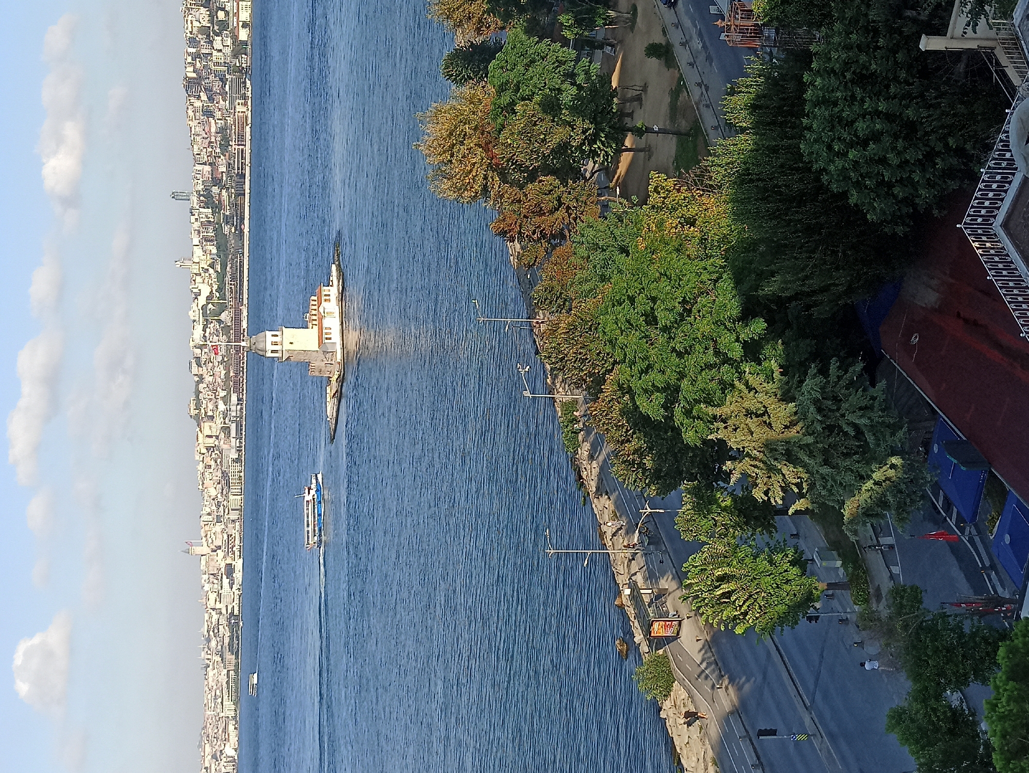 This is a photograph of the Maiden's Tower (Kız Kulesi) in Üsküdar, Istanbul, Turkey. The tower is situated on a small islet in the Bosphorus strait, with the Üsküdar coastline and the dense cityscape of Istanbul stretching across the horizon. In the foreground, there is a tree-lined promenade and a road with a few pedestrians and a bus stop. The water is calm with two ferries visible in the distance. The sky is partly cloudy, and the lighting suggests it is either early morning or late afternoon.