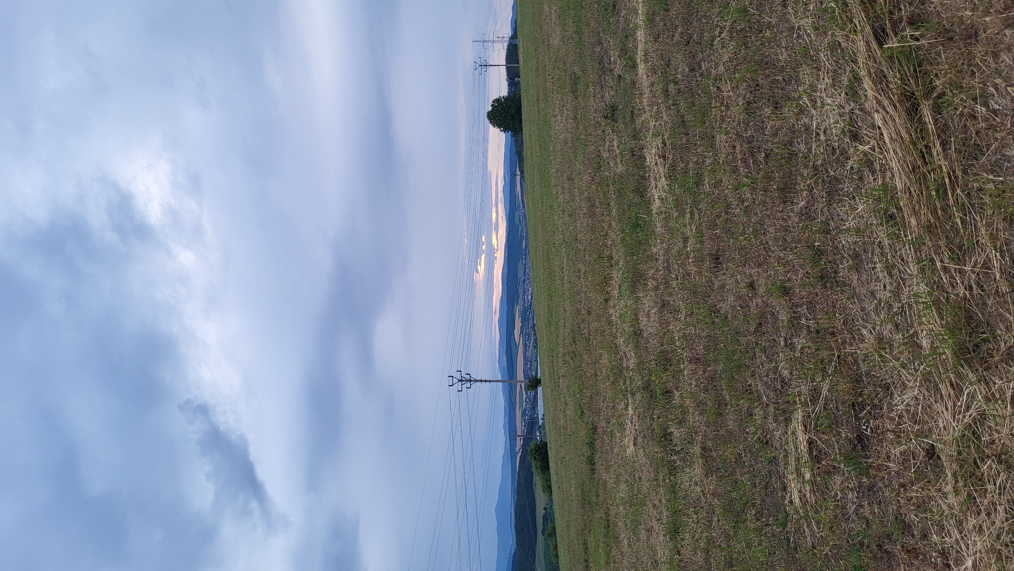 This is a vertically oriented, medium-distance, outdoor photograph taken in the city of Prešov, Slovakia. The foreground is dominated by a dry, brown, grassy field. In the mid-ground, a gentle slope leads to a village nestled in a valley, with rolling hills and mountains in the distance. Power lines are visible strung between utility poles across the landscape, and a few scattered trees dot the scene. The sky is overcast with dramatic clouds, but there is a sliver of sunlight breaking through near the horizon, indicating either dawn or dusk. The overall atmosphere is serene and rural.