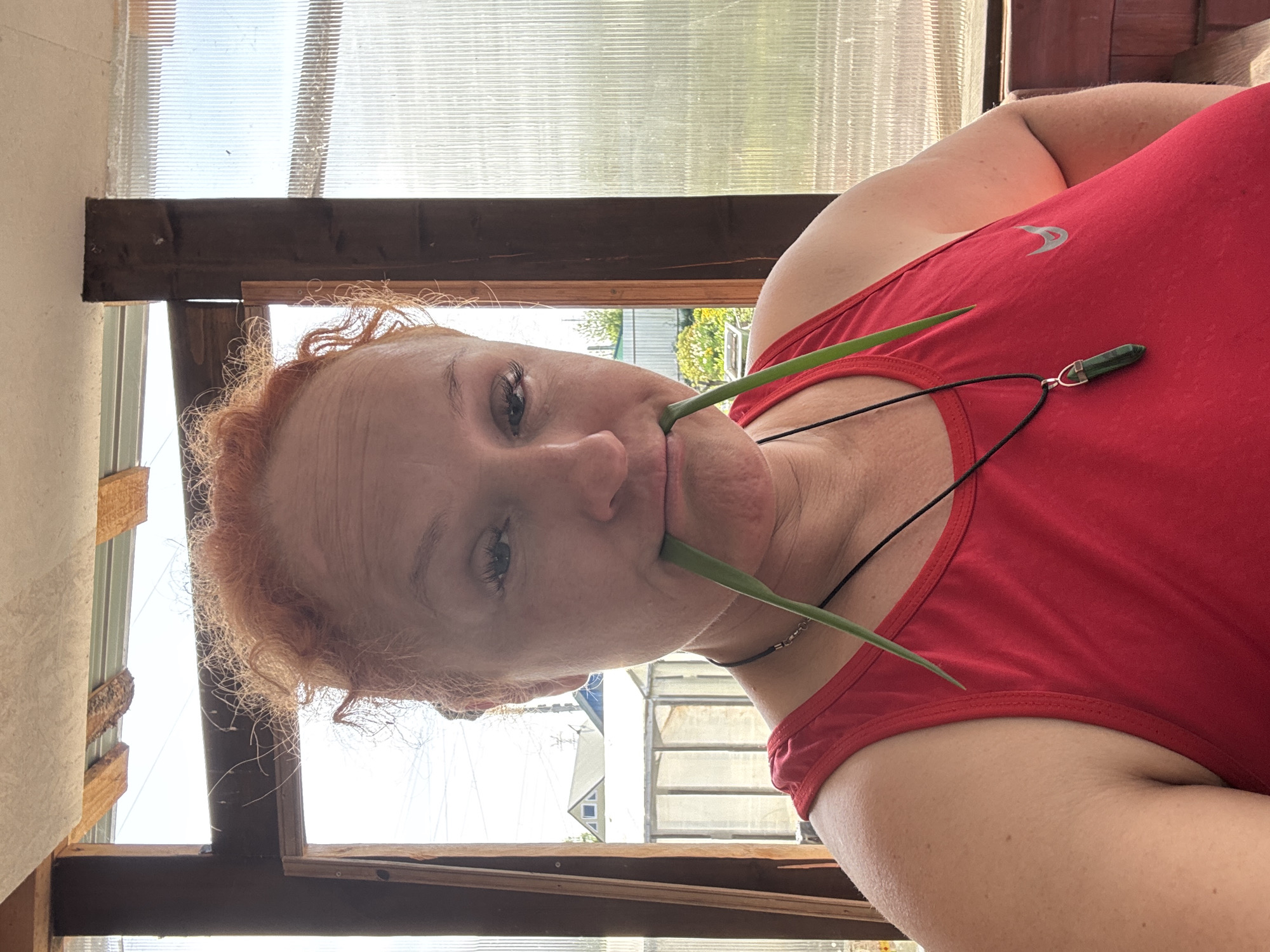 A woman with red hair is holding two green blades of grass in her mouth. She is wearing a red tank top and a black necklace with a green pendant. The location appears to be inside a structure with a translucent roof and wooden frames, possibly a greenhouse or sunroom. In the background, outside the structure, there are buildings and greenery, typical of a residential area. The city of Chaikovsky, Russia is not identifiable in the scene.
