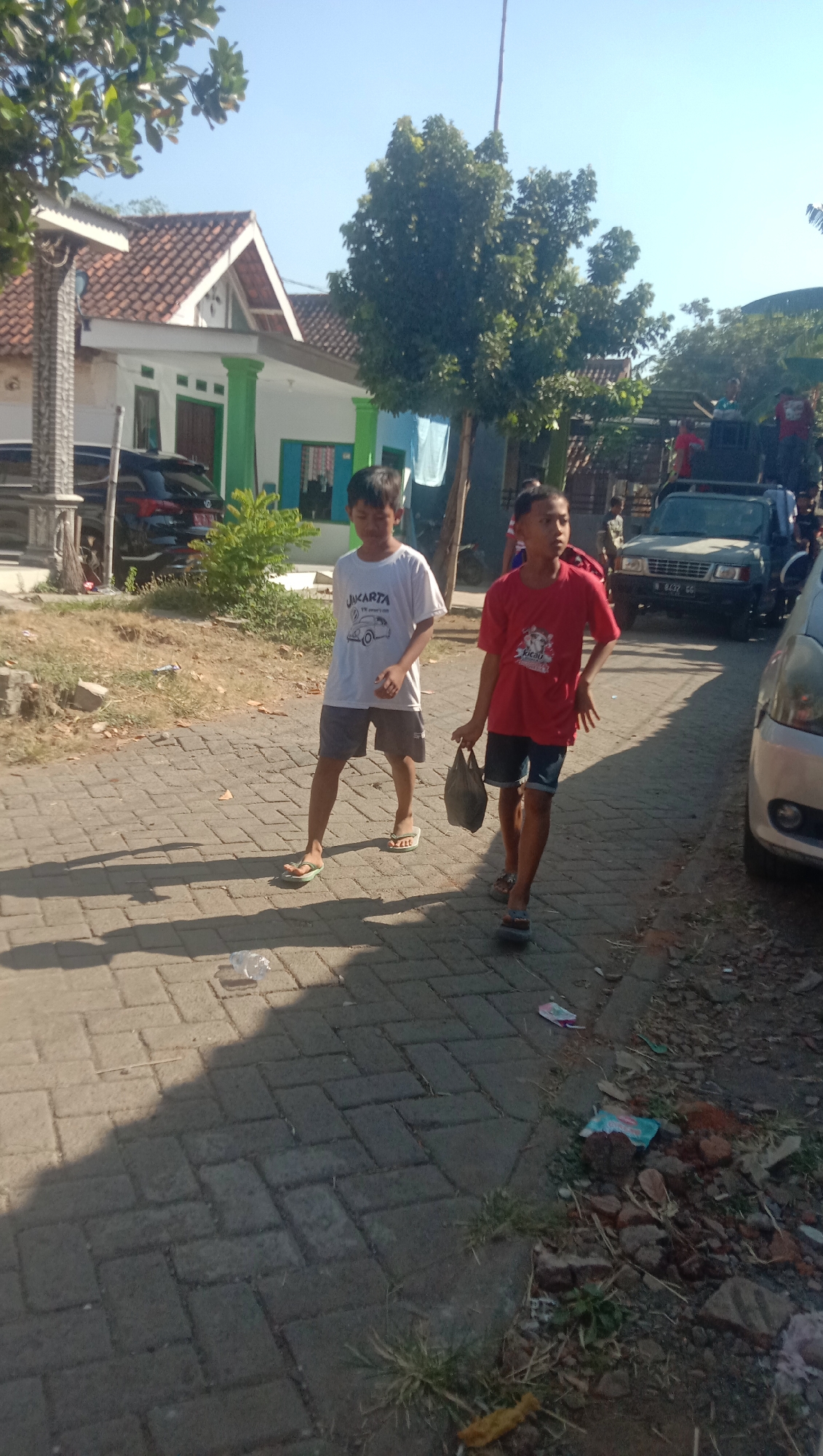 The image shows two young boys walking on a paved street in the city of Ngempit, Indonesia. The boy on the left is wearing a white t-shirt with the word "JAKARTA" printed on it, and grey shorts. The boy on the right is wearing a red t-shirt and blue shorts, and is carrying a dark-colored plastic bag. They are walking towards the right of the frame. In the background, there is a house with a white and green facade, trees, and a black car parked in front of the house. To the right, there is a silver car and a truck. The sky is clear and blue, indicating a sunny day. The ground is paved with interlocking bricks, and there is some debris on the side of the road, including a plastic bottle and some bricks. The overall scene suggests a casual, outdoor setting in a residential area.
