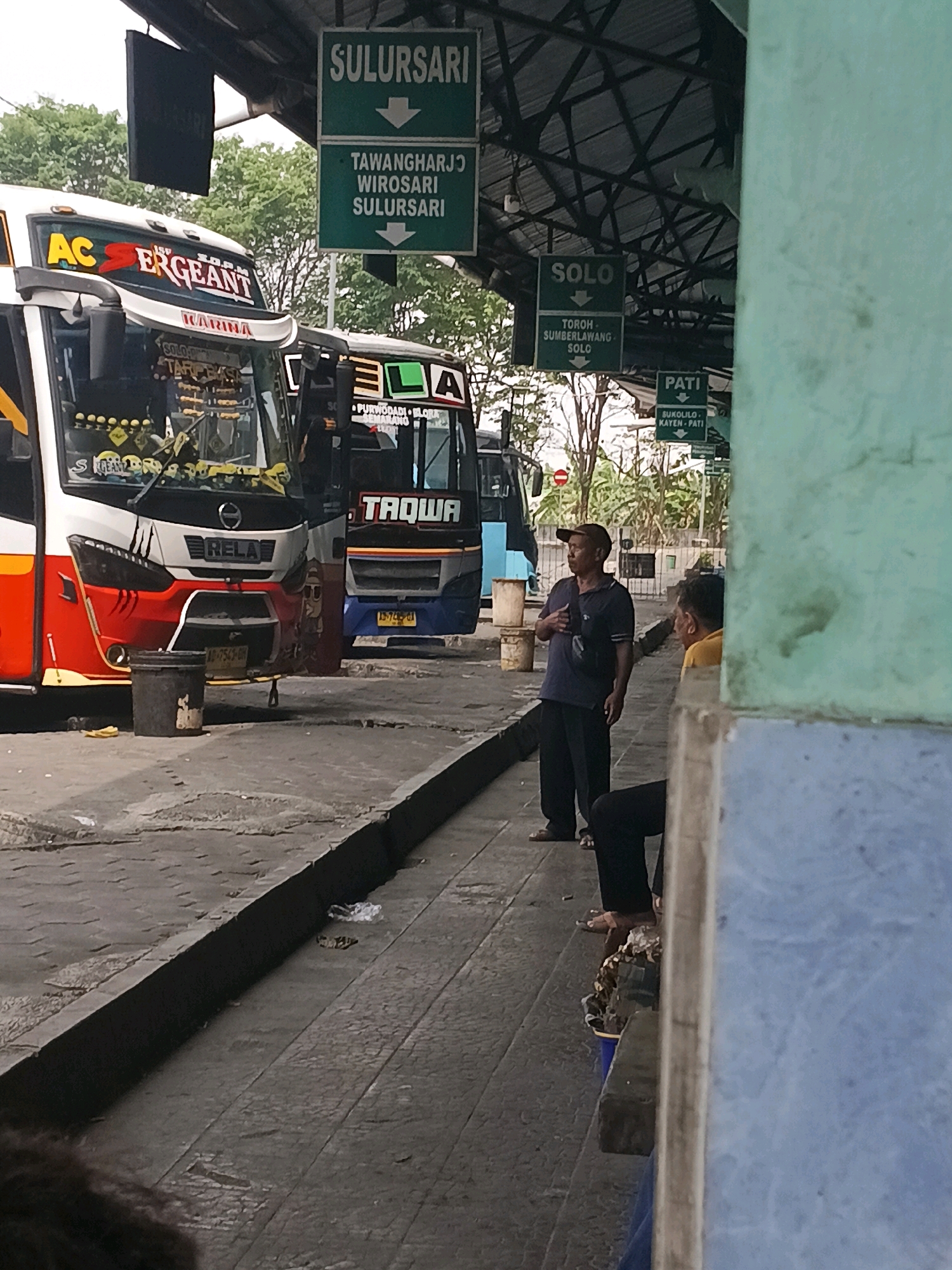 A photograph taken outdoors at a bus terminal in Kuripan, Indonesia, shows two buses parked at a covered bay. The bus on the left is white with red and yellow accents, and the bus on the right is blue and white. Both buses have signage indicating their destinations. Several green directional signs are visible, including one that reads "SULURSARI" and another for "TAWANGHARJO WIROSARI SULURSARI." Another sign reads "SOLO" with a directional arrow, and below it, "TOROH SUMBERLAWANG SOLO." A sign for "PATI" is also visible. Two men are standing near the bus on the right. One man, wearing a dark blue shirt and a baseball cap, stands facing left, with his hand in his pocket. Another man, wearing a yellow shirt, is seated to the right of the first man, partially obscured by a blue wall. The ground is paved, and a black trash can is visible near the front of the bus on the left. Trees are visible in the background.