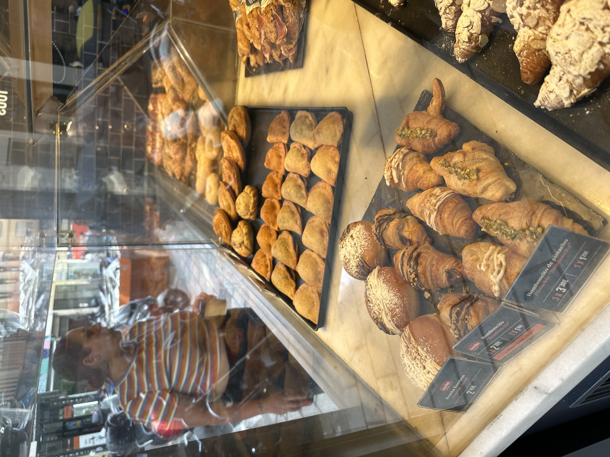 The image shows a vibrant display of pastries inside a glass case, likely in a bakery or cafe. The scene is viewed from an angled perspective, with reflections on the glass adding depth and revealing the surrounding environment and the person taking the photo.
Inside the display, various baked goods are arranged on black trays. On the left, a tray holds numerous golden-brown, possibly savory, turnovers or empanadas. To the right, another section showcases a variety of sweet croissants and pastries. Some croissants are generously dusted with powdered sugar and almond slivers, while others feature chopped pistachios as a topping. There are also pastries with chocolate drizzles or fillings.
A person's reflection is prominent on the left side of the glass. They are wearing a distinctive striped shirt with orange, blue, white, and yellow/gold stripes, and appear to be observing or photographing the display. The reflection also shows glimpses of the shop's interior, including what looks like a brick pattern wall and other fixtures.
Visible text on price tags provides details about the items. One tag reads "nocilla Bomba Bombón 245€", indicating a chocolate bomb pastry. Another tag for "Bomboncito" offers pricing options: "3X 280€" and "1X 100€", with a note "Ya última hora" (last hour), suggesting a special offer. A third tag is for "Bomboncito de pistacho Pistachio 'Bomboncito'", priced at "3X 330€" and "1X 120€". A partial reflection of "1003" is also visible on the glass. The overall scene depicts a well-stocked and inviting pastry display.