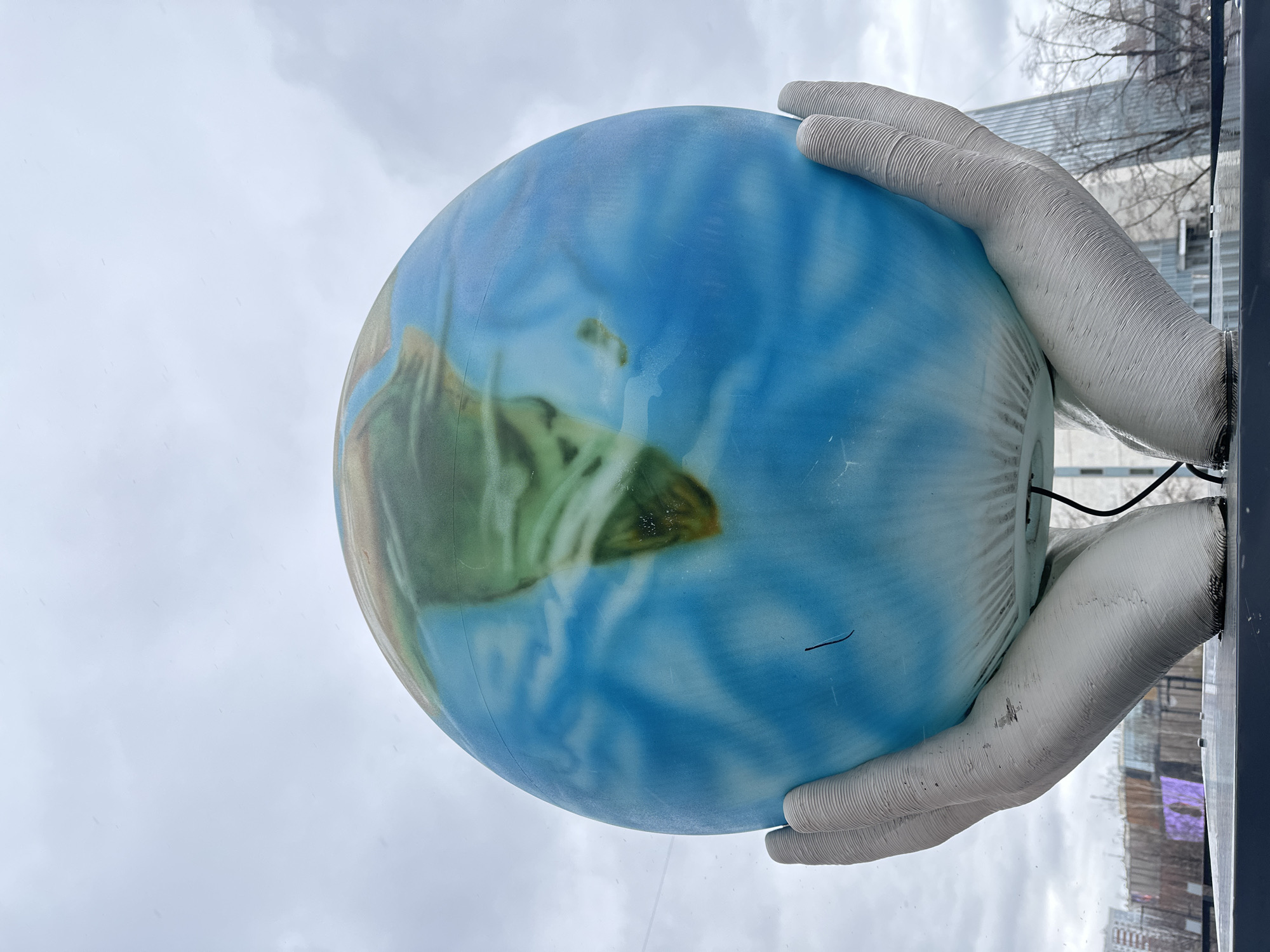 A medium, angled shot features a globe held by two large, white hands, likely an art installation. The globe is painted with blue oceans and green landmasses. The hands are textured with a ridged pattern, resembling inflated tubes. A black cable extends from the base of the installation. The background reveals an urban setting with buildings, bare trees, and an overcast sky. The scene is located in the city of Perm, Russia. The globe and hands are positioned near a window.