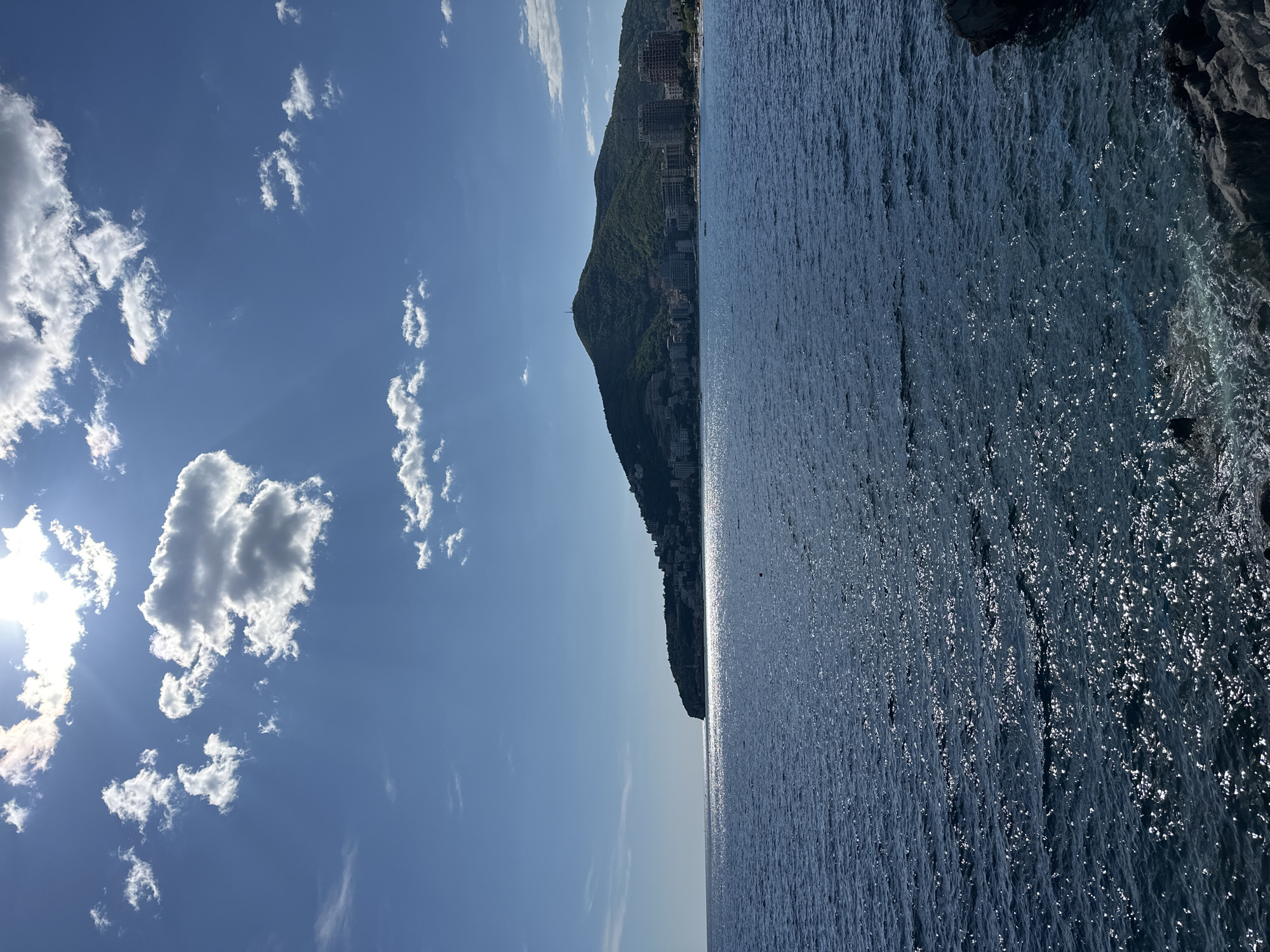 The image displays a picturesque coastal landscape in Budva, Montenegro, under a bright, sunny sky. The scene is bisected by a prominent, dark green mountain or hill covered in lush vegetation, which rises from the sparkling blue sea. Numerous buildings, likely hotels or residential complexes, are visible nestled into the mountain's slopes, particularly along its base and higher up, showcasing urban development integrated with the natural terrain.
The sky, occupying the upper left quadrant, is a clear, vibrant blue dotted with scattered white cumulus clouds. The sun, bright and partially hidden by a cloud on the far left, emits discernible rays, illuminating the sky.
The sea, filling the right side and foreground of the image, is a deep, rich blue. Its surface glistens with countless reflections of sunlight, indicating gentle ripples or small waves. In the far foreground on the right, dark, rugged rock formations meet the water's edge, where some white foam suggests the interaction of waves with the shore.
The time of day is clearly daytime, likely late morning or early afternoon, given the bright illumination and high sun. The weather is sunny with some scattered clouds. No people or visible text are present in the image. The overall impression is one of serene natural beauty combined with coastal development.