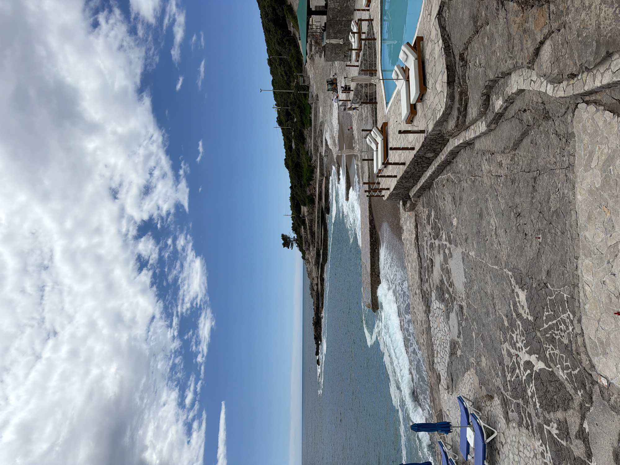 The image depicts a coastal scene in Prijevor, Montenegro. The view is taken from an elevated position. The left side of the image features a sky with white, billowy clouds against a blue background. Below the sky is a view of the sea, extending to the horizon. Waves are crashing against the rocky shoreline.
To the right is a rocky outcrop with visible veins of white. On the outcrop, there are beach chairs and umbrellas. Further inland, a swimming pool and sun loungers are visible. A line of green vegetation runs along the top of the cliff.