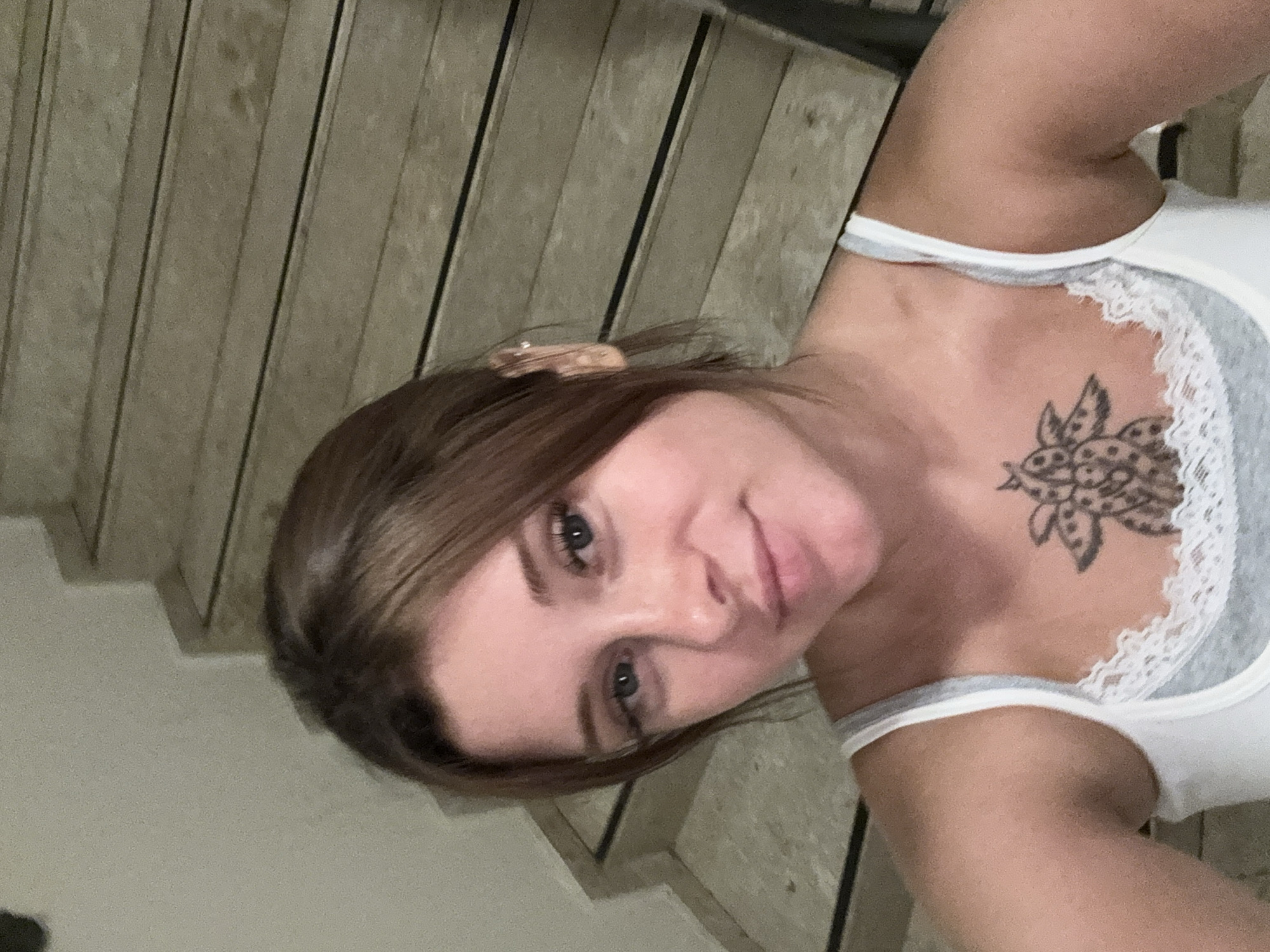 A young woman with light brown hair and blue eyes is taking a selfie on a set of stairs. She is wearing a white tank top with grey trim and lace detailing. She has a tattoo on her chest. The stairs are made of wood with dark trim and are located indoors. The image is a close-up of the woman's face and upper body.