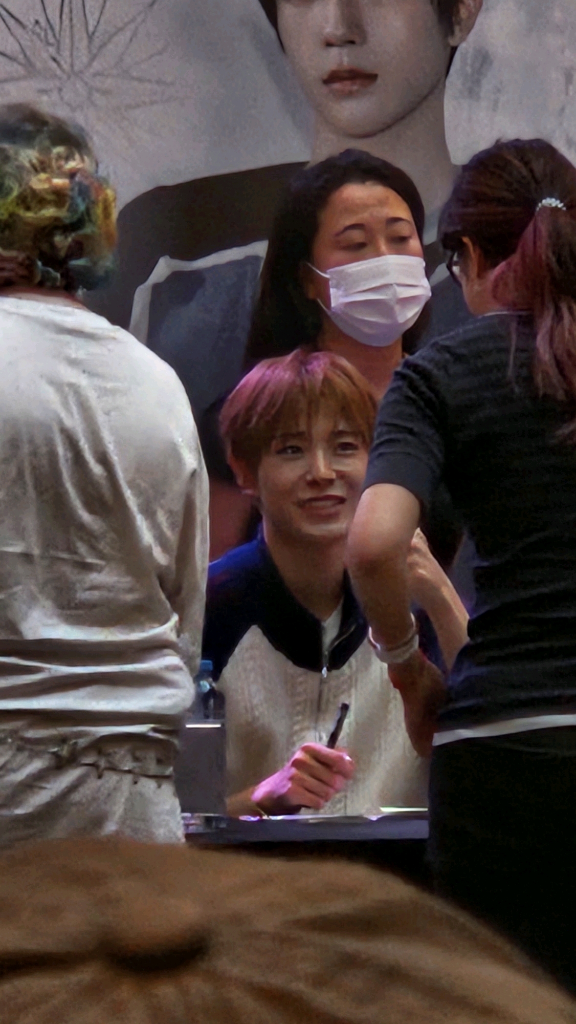The image captures a candid moment likely during a fan event or autograph session in Barcelona, Spain. In the foreground, a person with vibrantly colored, textured hair is partially visible from the back, wearing a loose-fitting, light-colored garment. To their right, a young male with reddish-brown hair is smiling and looking towards someone off-camera, possibly holding a pen. He is wearing a jacket with a white front and dark collar and sleeves. Behind him, a woman wearing a white face mask is looking downwards, and further to the right, another person with their back to the camera, wearing glasses and with dark hair tied back, appears to be interacting with someone. The background features a large graphic or poster of a person's face, suggesting a promotional event. The overall atmosphere appears to be one of excitement and personal interaction.