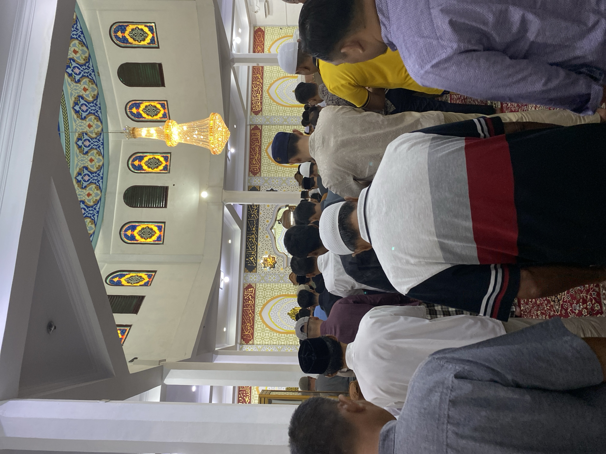In the city of Lhokseumawe, Indonesia, a group of men are gathered in a mosque, facing the front for prayer. The interior of the mosque is ornately decorated, with intricate patterns on the ceiling and walls, featuring Arabic calligraphy and geometric designs. A large, ornate chandelier hangs from the ceiling, casting a warm light on the scene. Stained-glass windows with colorful patterns are visible in the upper sections of the walls. The men are dressed in various traditional and casual attire, including prayer caps. The floor is covered with a red carpet with intricate patterns. The focus is on the backs of the men as they stand in prayer, creating a sense of community and devotion.