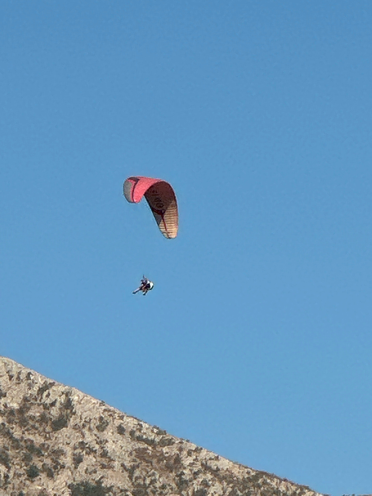 A paraglider is in flight against a clear blue sky. The paraglider, with a pink and tan canopy, is suspended in the air, with a pilot visible below. In the foreground, the rocky, sparsely vegetated slope of a mountain rises from the bottom of the frame. The scene is set in Boreti, Montenegro, a location known for its coastal mountainous terrain.