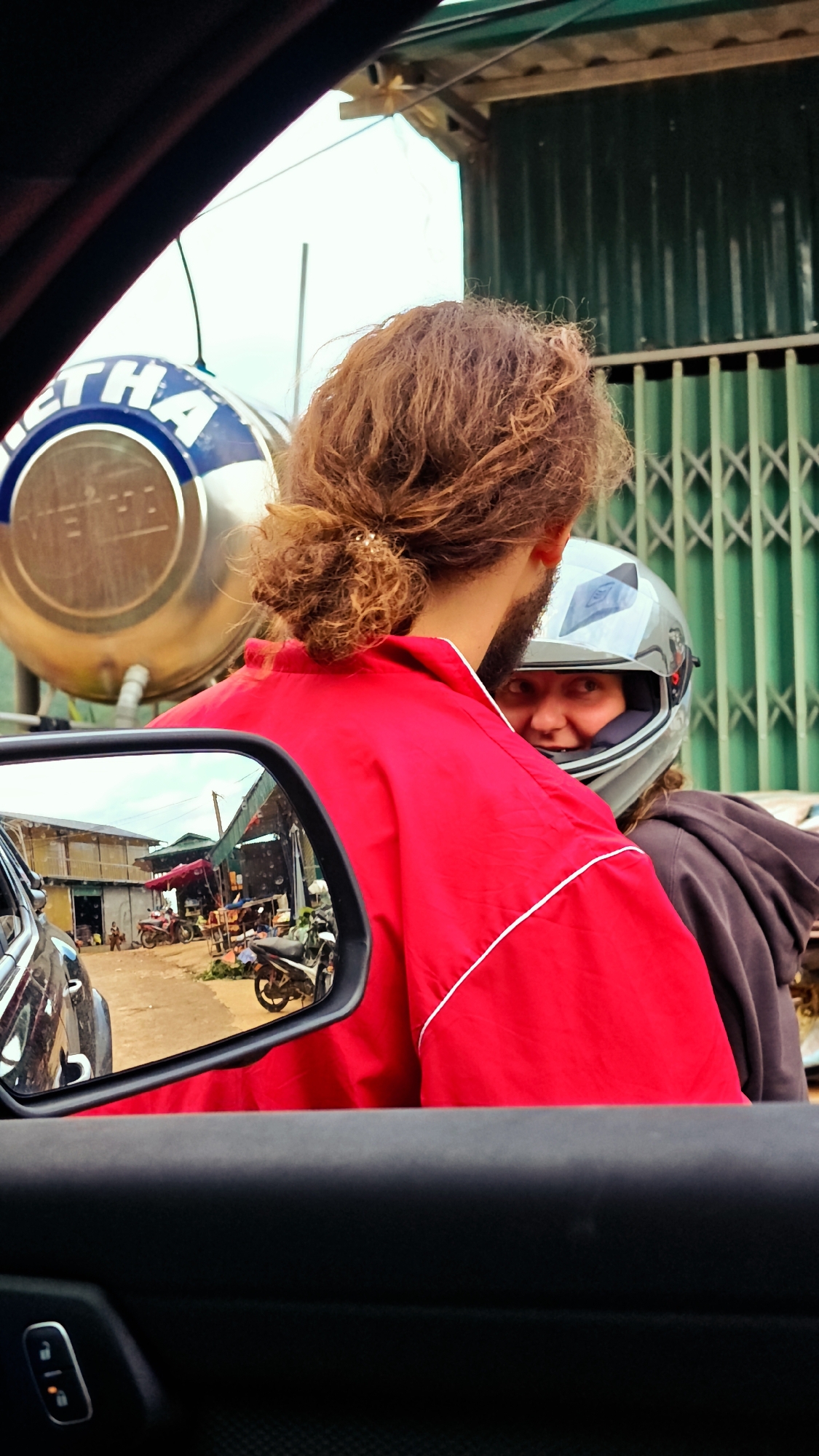 The image, captured from the interior of a vehicle in Xín Vàng, Vietnam, presents a street scene with two individuals on a motorbike as the central focus. The perspective is from the passenger side, with the dark gray car door panel and side mirror occupying the foreground. A silver control panel with two illuminated icons (likely door lock and window control) is visible on the lower left of the door panel.
Outside the window, a person with curly, reddish-brown hair tied in a bun is seen from behind, wearing a bright red long-sleeved jacket with a thin white vertical stripe on the side. A dark beard is visible along their jawline. Positioned behind this individual on the motorbike, a second person wears a light grey full-face motorcycle helmet with a dark visor. Their face, partially visible through the helmet's opening, shows a slight smile. This individual is wearing a dark brown or black hooded garment.
In the upper left background, a large, metallic, cylindrical tank is visible, featuring blue lettering "ETHA" or "IETHA" on a white background, indicative of a local brand. To the right, a dark green corrugated metal building facade rises, with a lighter green metal accordion-style security gate visible below it. The sky above is bright and overcast.
The car's side mirror reflects a detailed streetscape of Xín Vàng. A dirt or unpaved road is visible, flanked by simple, somewhat rustic buildings or market stalls, some with corrugated metal roofs and exposed structural elements. Several motorbikes are parked or appear to be navigating the street. Market goods or general clutter are discernible outside the buildings, and faint figures of people are present in the distance. One structure prominently features a red awning.