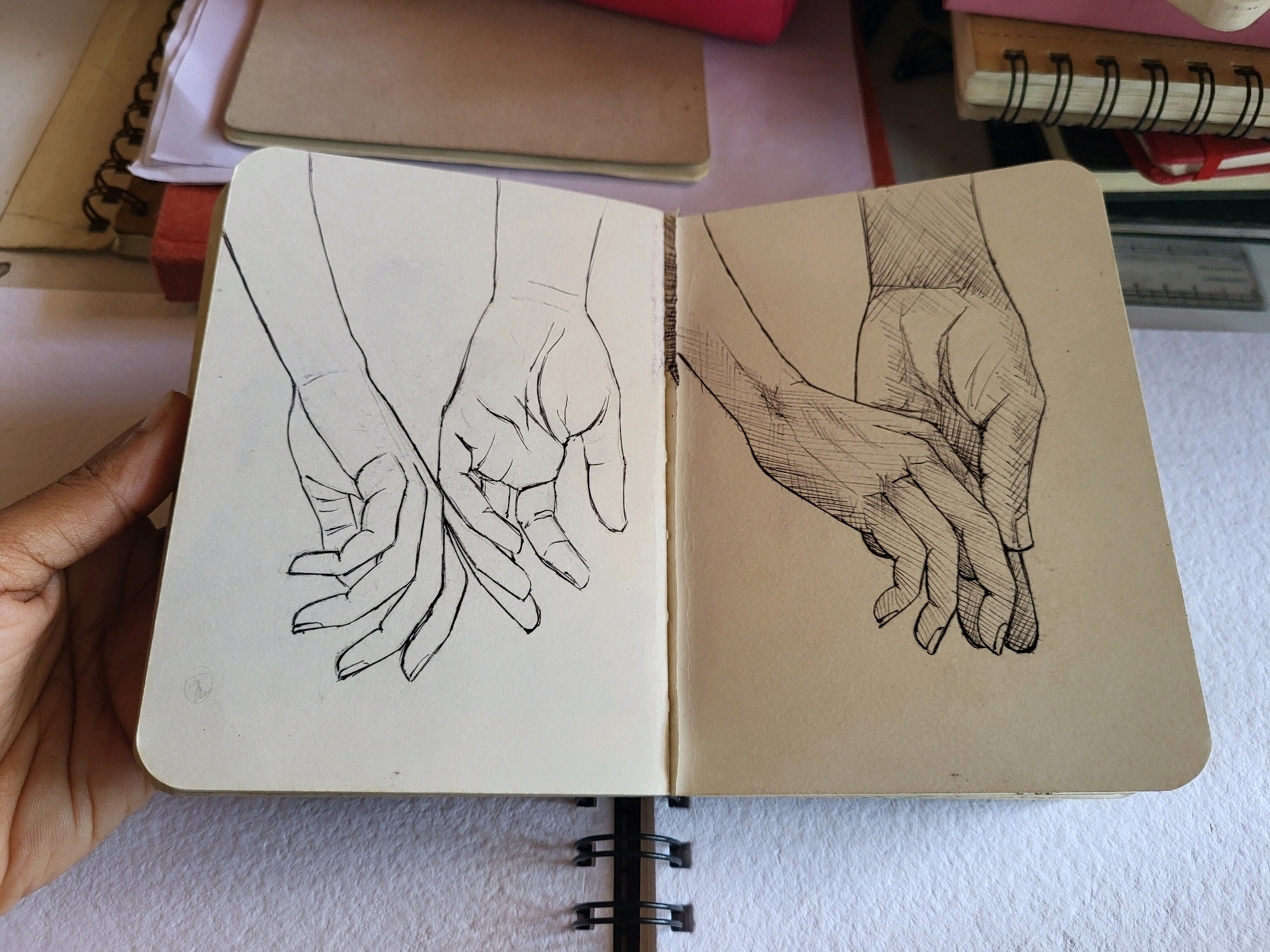The image shows an open sketchbook with two ink drawings of clasped hands. The left page is blank paper and features a line drawing of two hands. The right page is on tan paper and shows a more shaded drawing of two hands. The sketchbook is lying on a white textured surface, likely a table. In the background, stacked notebooks and papers are visible. The lighting suggests an indoor environment. There is no information or visual cue within the image to suggest the location is Koduvayur, India, or any specific city.