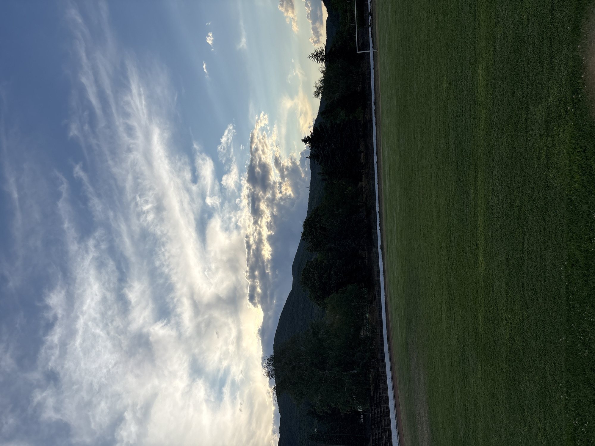 The image shows an outdoor view of a green grass field, possibly a sports ground, with a track or path behind it. Beyond the field, there are tiered seating structures, followed by a line of dark green trees and then a mountain range. The sky above is a mix of blue and white clouds, with sunlight peeking through some of the darker clouds near the mountain. A white goalpost is visible on the right side of the field. The scene is captured during what appears to be late afternoon or early evening, given the lighting and the clouds. The location is identified as the sports ground in Boboshevo, Bulgaria. There are no people or animals visible in the image.