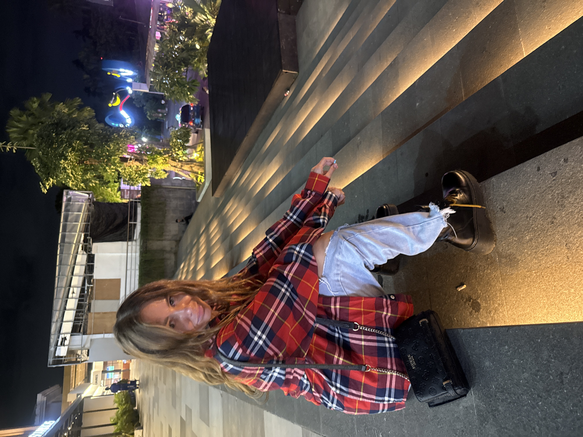 At night in what appears to be Kuta, Indonesia, a woman sits on gray stone stairs. The stairs have integrated strip lighting that casts a yellow glow upward. She is wearing a red and blue plaid shirt, light blue jeans, and black platform shoes with yellow shoelaces. A black Guess purse with a silver chain rests near her. Street lights and buildings are visible in the background, with a colorful amusement structure situated among trees. A cigarette butt lies on the ground near her feet.