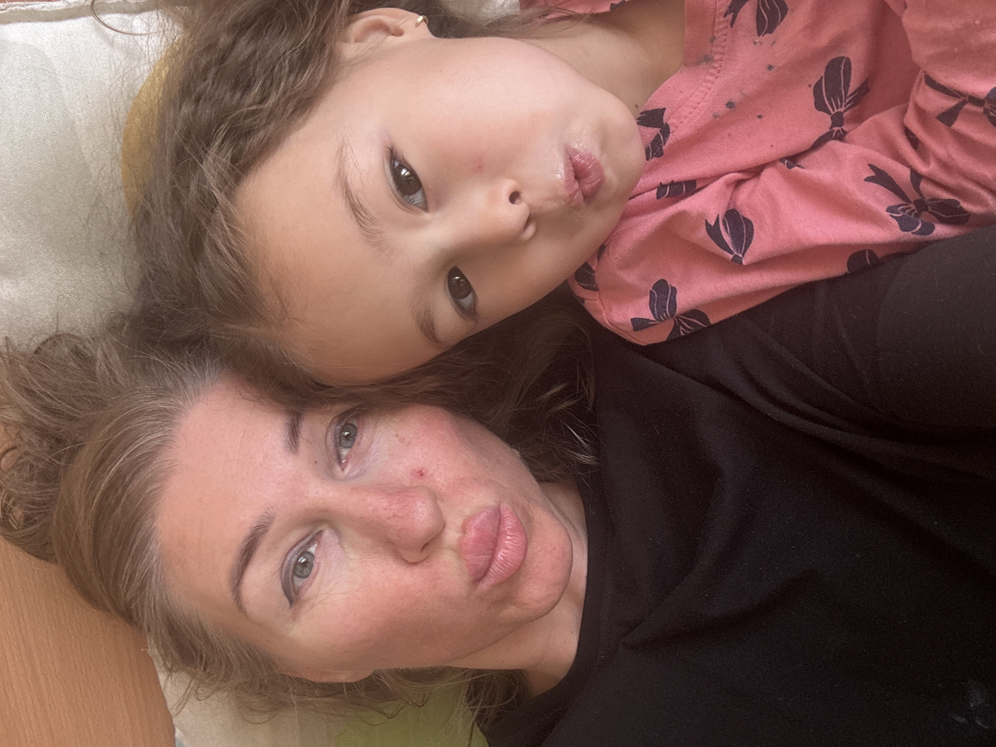 Two individuals, likely a mother and daughter, are positioned close together, possibly lying down. The child is wearing a pink top with dark blue bow patterns and has brown hair. The adult is wearing a black top. Both are facing the camera with pursed lips. The background is indistinct, but suggests an interior setting. There is no clear indication of location, and the claim that it is Tashkent, Uzbekistan, cannot be verified.