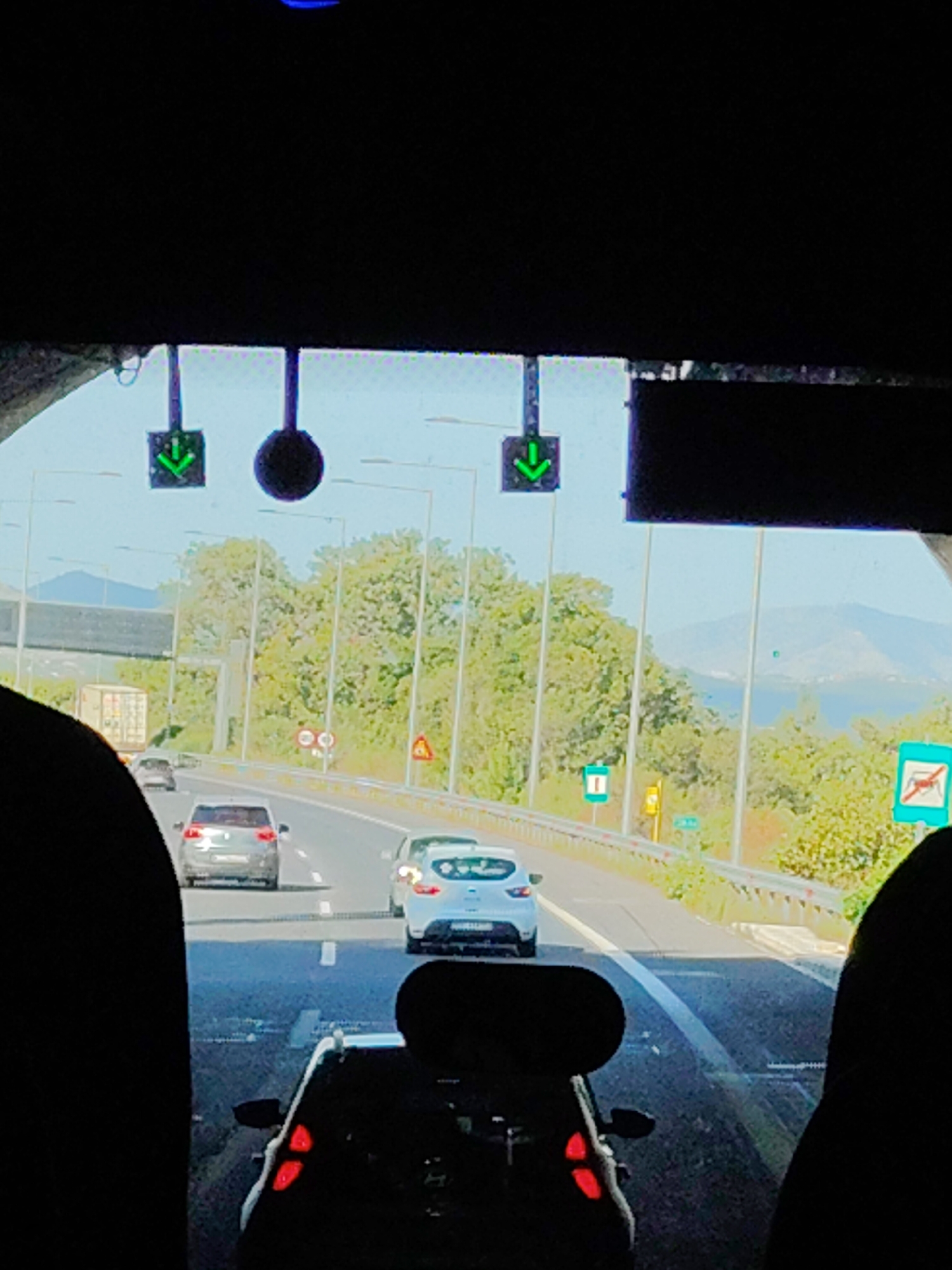 The image shows a view from inside a vehicle exiting a tunnel onto a highway. The time of day appears to be daytime, with bright sunlight visible. Two green arrows pointing downwards are displayed on a gantry above the highway, indicating the direction of travel. A large, dark, circular object, possibly a light or a sign, is also present on the gantry to the right of the arrows.
Several cars are on the highway, including a gray hatchback in the left lane and a white hatchback in the right lane. A truck with a white trailer is visible in the distance on the left side of the highway. Road signs and streetlights line the highway.
The landscape beyond the highway includes green trees and hills in the distance, with a body of water and mountains visible under a clear blue sky. The right edge of the image shows the silhouette of a person or object in the foreground, obscuring part of the view.
The presence of signs indicating a highway, traffic signals, and road infrastructure suggests this is a public road. The overall scene depicts normal traffic flow on a sunny day. The Municipal Unit of Ampelakia is located in the region of Attica, Greece, and this highway is likely part of the Greek national road network.