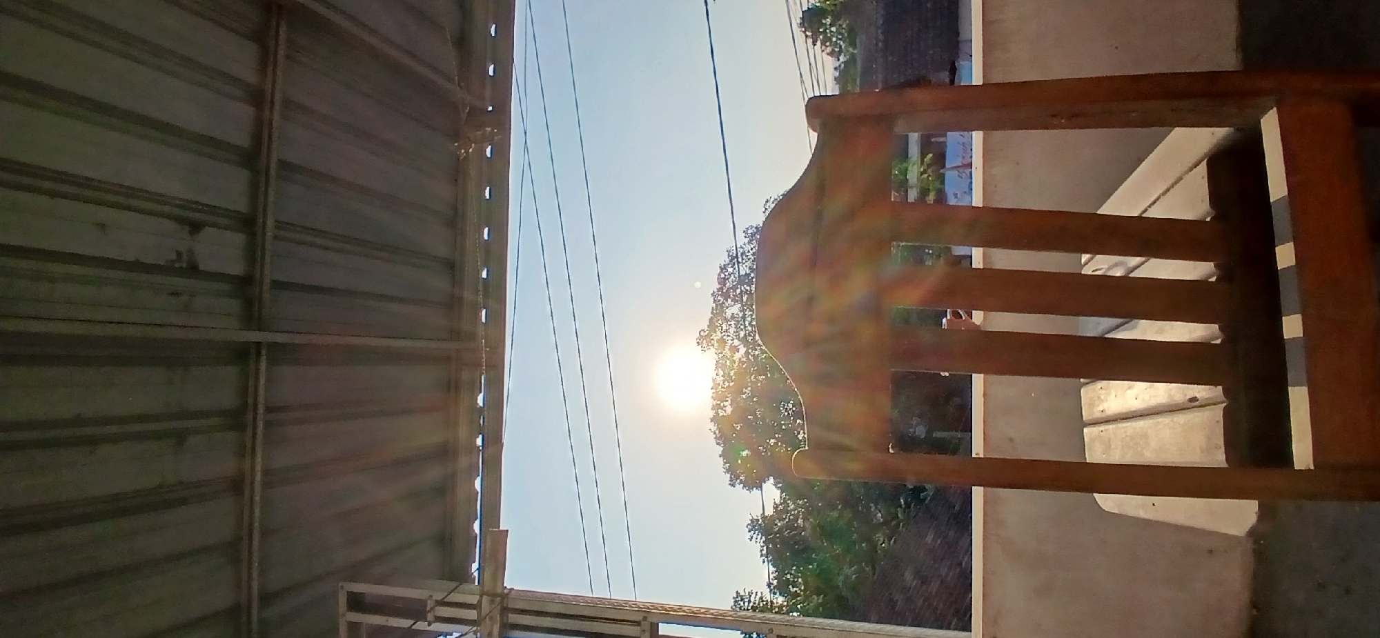 A wooden chair faces a bright sun on a clear day, with a corrugated metal roof overhead. The scene is set on a balcony or porch, overlooking trees and rooftops in Kademangan Kulon, Indonesia. The sun's rays create a lens flare effect, illuminating the chair and the sky. Power lines are visible across the sky, suggesting an urban or semi-urban environment. The overall impression is one of a serene, sun-drenched afternoon.