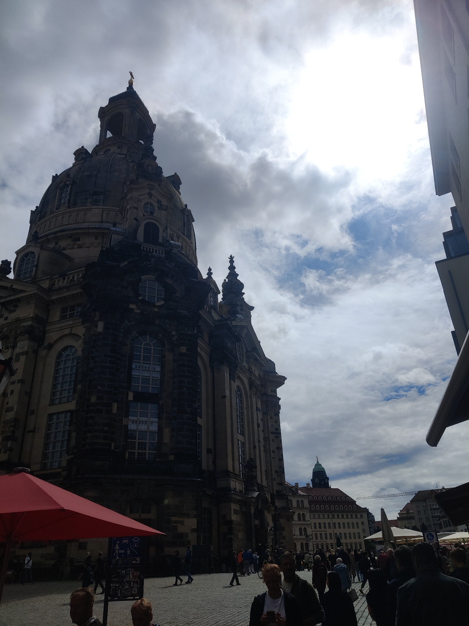 The image shows the Frauenkirche, a Lutheran church in Dresden, Germany, on a cloudy day. The church's dome and spire are visible against a dramatic sky. In the foreground, people are walking on a cobblestone plaza, some are sitting at tables under a red umbrella. The architecture of the church is baroque, with intricate detailing and large arched windows. Another building with a green dome is visible in the distance. The sun is partially obscured by clouds, casting a diffused light on the scene.