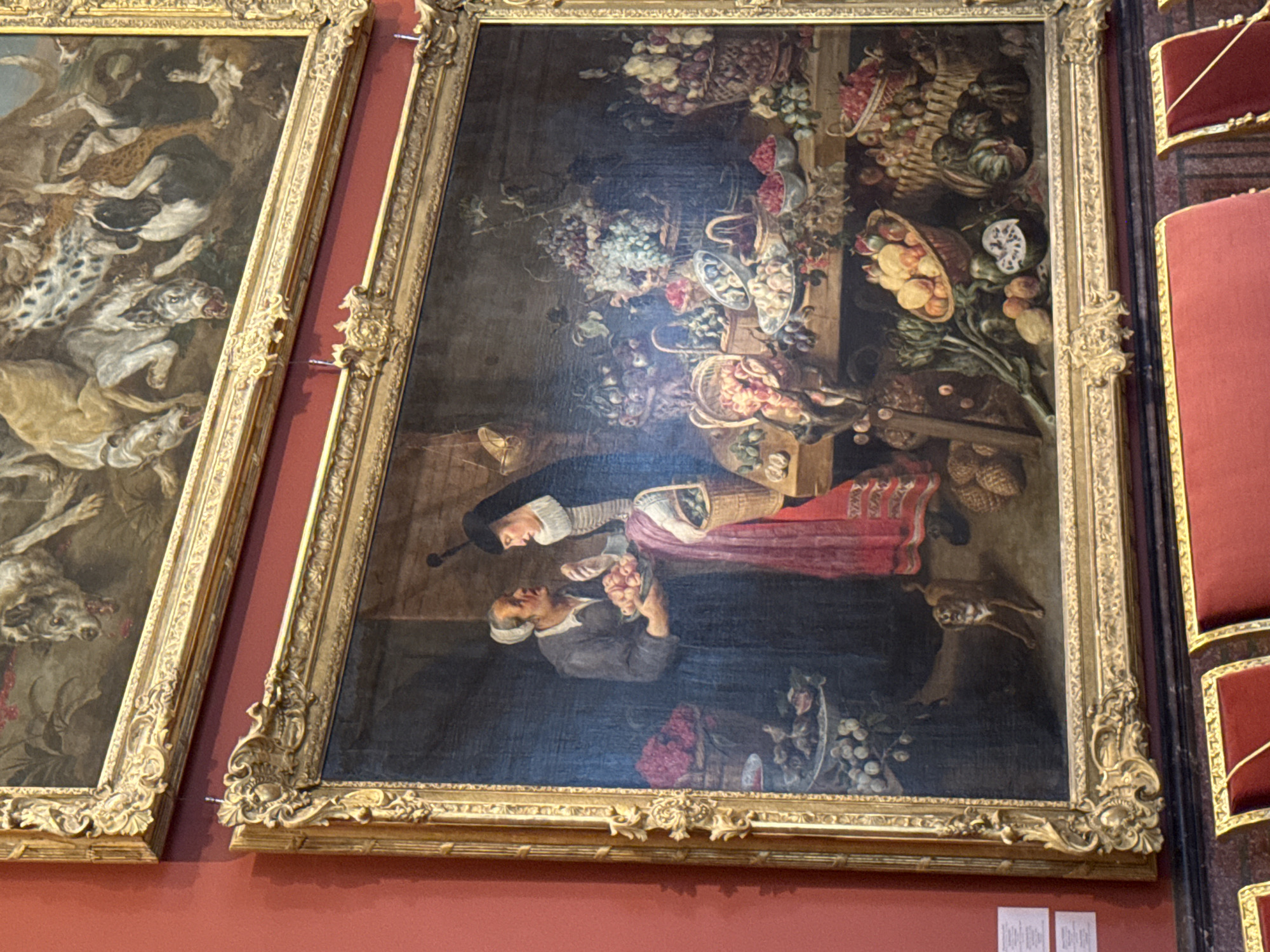 The image shows two paintings hanging on a red wall. The lower painting is a detailed still life and genre scene, framed in ornate gold. It depicts two women interacting in what appears to be a marketplace or domestic setting, surrounded by abundant displays of fruits and vegetables. The woman on the left, wearing a dark dress and white cap, holds a platter of light-colored fruits, possibly apples or pears, and offers them to the woman on the right. The second woman, dressed in darker attire with a distinctive white collar and black headdress, is looking at the fruit. The foreground is laden with baskets and bowls overflowing with various fruits like grapes, pomegranates, peaches, and what appear to be gourds or pumpkins. A small dog stands on its hind legs in the lower left of the painting, looking towards the women. The upper painting, also in a gold frame, is a hunting scene featuring a pack of dogs in various poses of aggression, with some appearing to be tearing at something indistinct. Both paintings are typical of Baroque or Rococo art. The presence of museum benches in red velvet in the foreground suggests this is an indoor exhibition space, likely a museum. Considering the style of art, the ornate frames, and the typical presentation of such works in grand institutions, this scene is likely located in the Hermitage Museum in Saint Petersburg, Russia, a world-renowned repository of art.