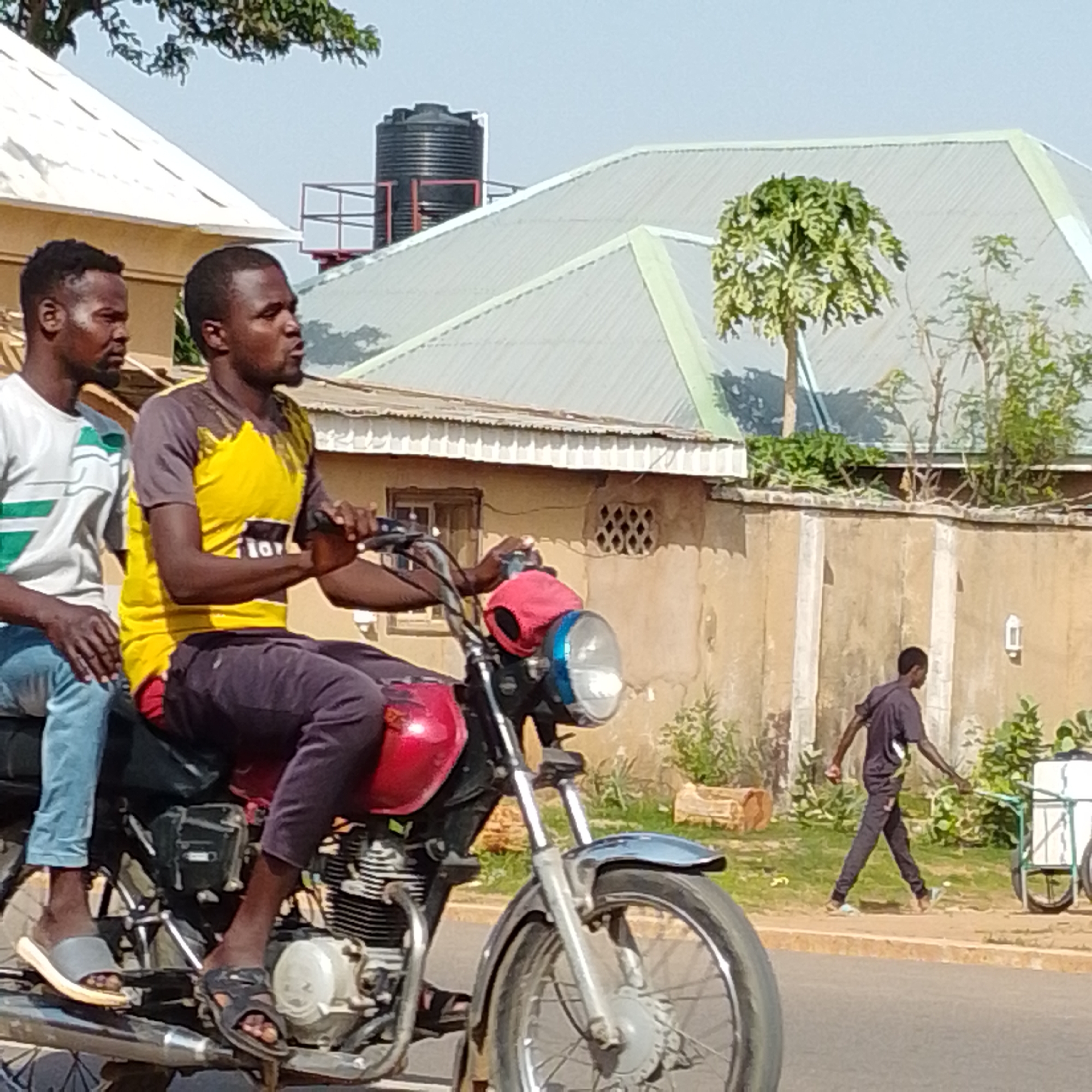In Gombe, Nigeria, two individuals are riding a motorcycle on a paved road. The person in front is driving, wearing a yellow shirt with black and white accents and dark trousers. The passenger behind him wears a white t-shirt with green stripes and light blue jeans. The motorcycle is red and black, with visible engine parts. In the background, a man is walking on a sidewalk, pushing a cart with a white container. The scene is set against a backdrop of buildings with corrugated metal roofs and stucco walls. Trees and vegetation are present, and a large black water tank is visible on top of one of the buildings. The sky is clear and bright.