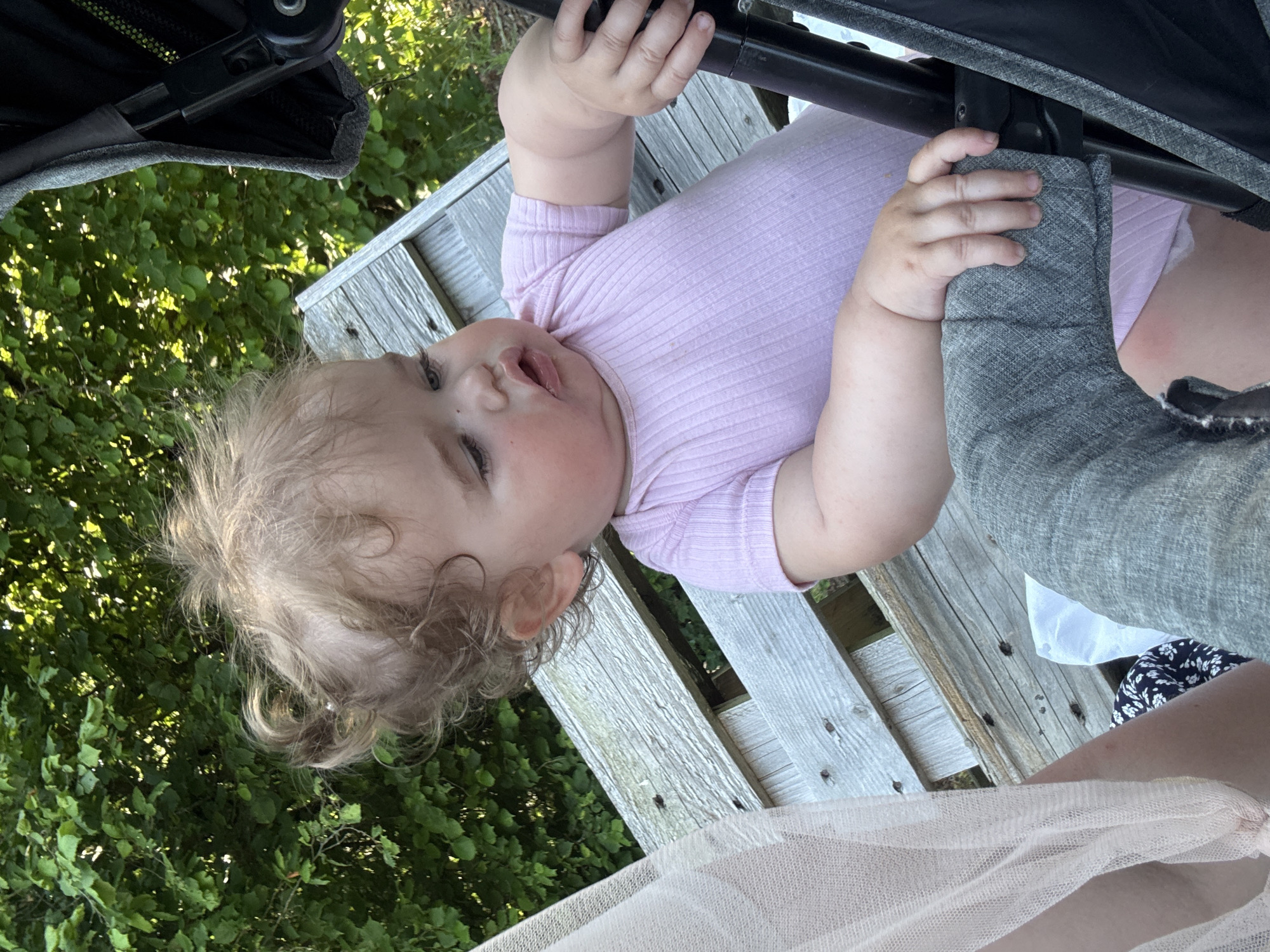 In Beltershain, Germany, a young child with curly blonde hair sits on a wooden bench, possibly part of a park or outdoor seating area, near dense green foliage. The child, wearing a pink onesie, is looking to the right of the frame, with its mouth slightly open. The child's left hand is resting on a gray padded bar, likely from a stroller or pram, and its right hand is also visible, gripping the same bar. A portion of light pink sheer fabric is visible on the left side of the image, and another person's legs, clad in patterned fabric, are partially visible below the child. The overall scene suggests an outdoor, possibly recreational setting during daylight hours.