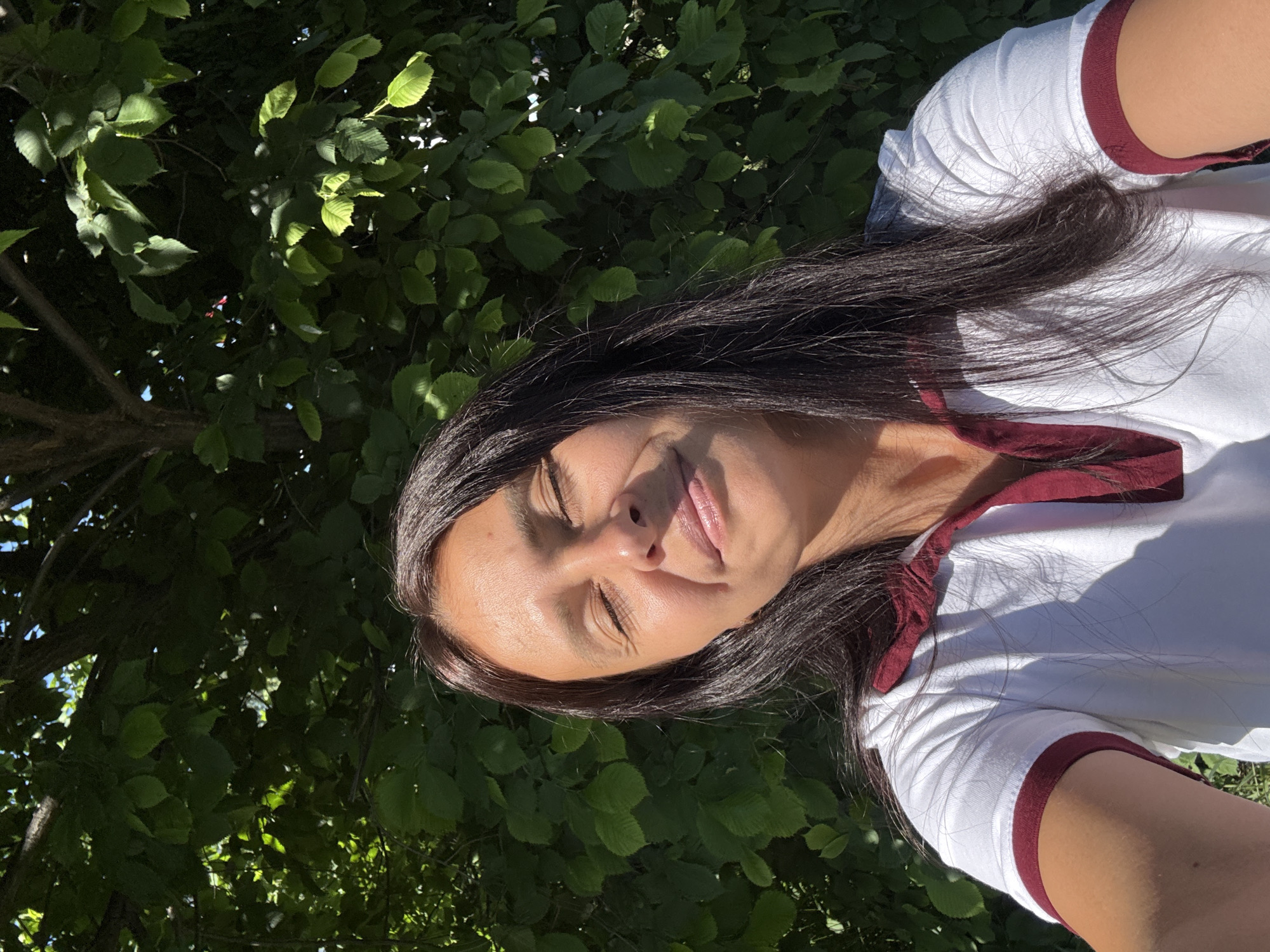 A woman is posing for a selfie outdoors in front of a dense green foliage background. She has long, dark hair and her eyes are closed, with a slight smile on her face. She is wearing a white polo shirt with maroon trim around the collar and sleeves. The sunlight is casting shadows on her face and the foliage. The image appears to be taken during the daytime. The location is not specified in the image or the provided information.