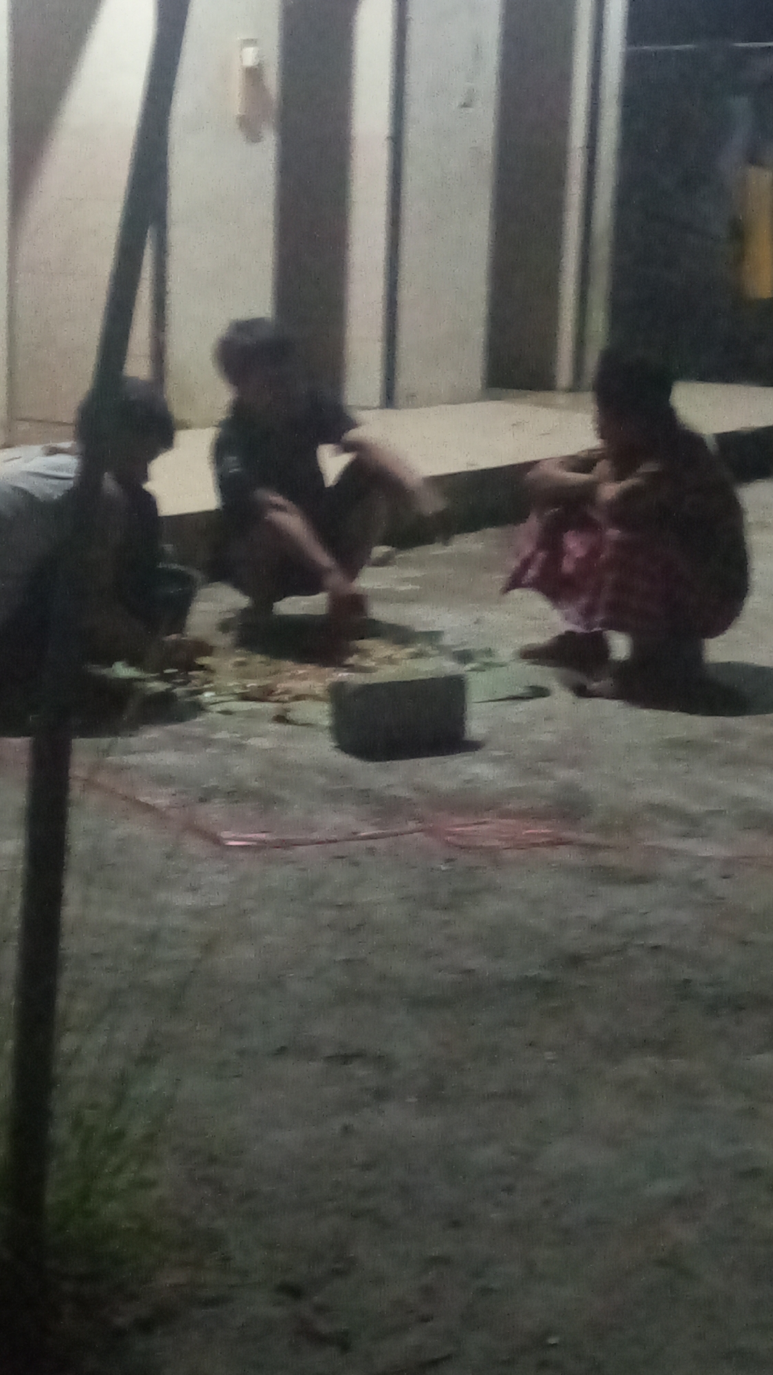 Three individuals are crouched on a paved outdoor surface, possibly in a public area or courtyard. The ground is a rough, grey concrete. In the center of the scene, there's a collection of items, including what appears to be fragmented debris and a dark, rectangular object resembling a brick or stone block. Red or pink thin rope-like material is visible on the ground in the foreground. Behind the individuals, there is a wall with vertical elements, suggesting a building or structure. The lighting is dim, indicating it may be evening or nighttime. The overall impression is one of a casual gathering or activity taking place outdoors.