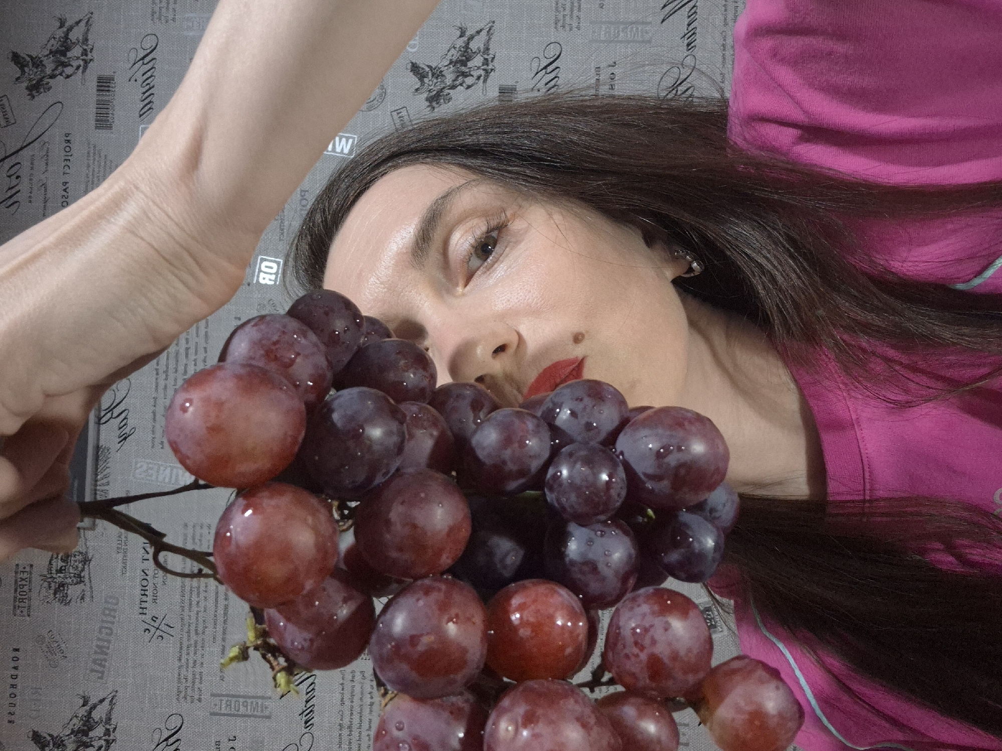 The image shows a woman with dark hair, red lipstick, and a mole on her face. She is holding a bunch of red and purple grapes. She is wearing a pink shirt. The background is a gray fabric with a newspaper print pattern.