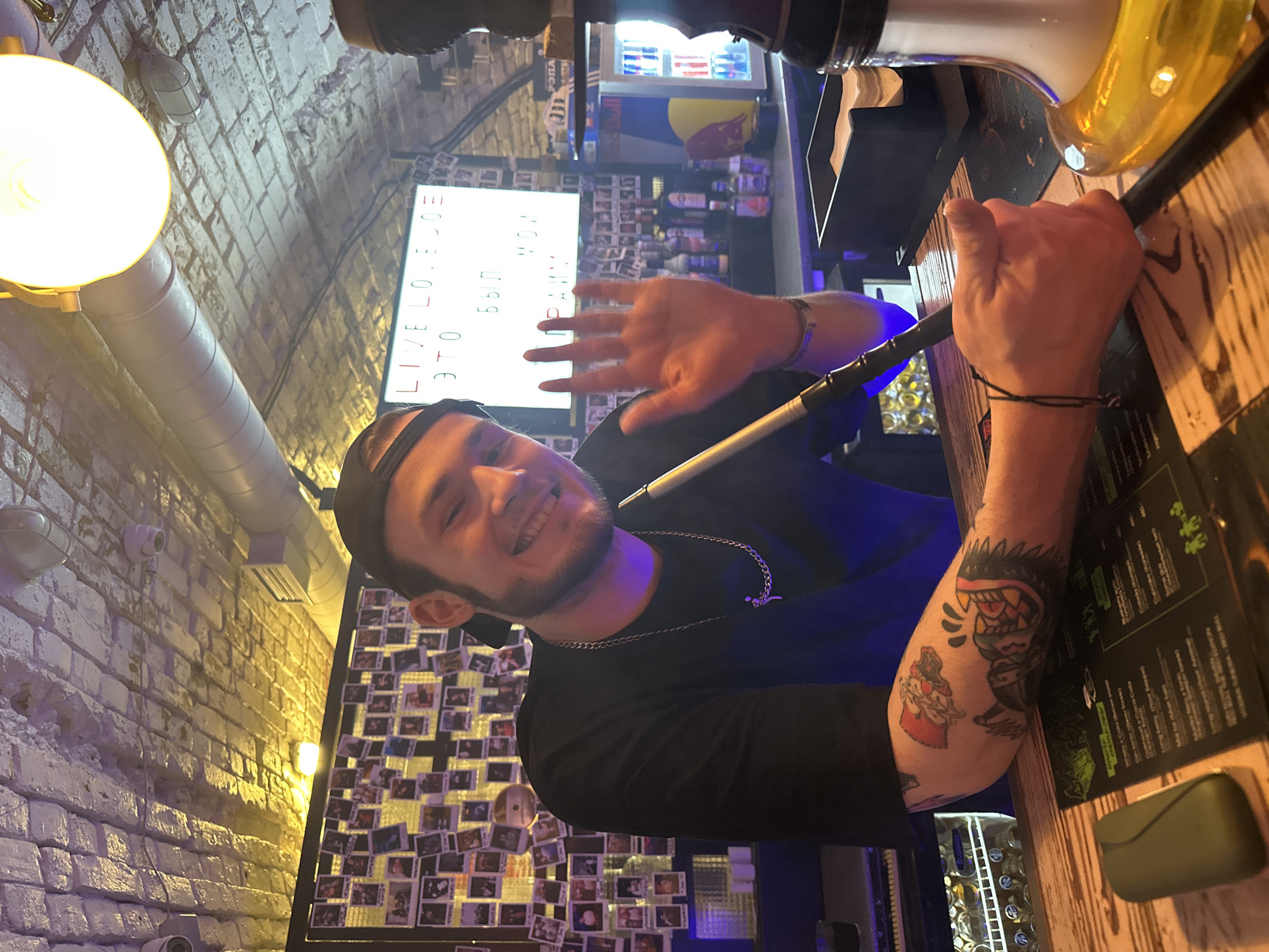 A young man with a beard and a black cap smiles broadly at the camera, posing indoors, likely in a bar or lounge in Krasnoyarsk, Russia. He is wearing a black long-sleeved shirt and a silver chain necklace. His right arm, adorned with several tattoos including a cartoonish monster-like creature and another figure, is bent, and his hand holds the hose of a hookah. His left hand is raised in a waving gesture, partially obscuring his face. The scene is dimly lit, suggesting an evening or night setting.
Behind the man, a brick wall is visible, along with industrial-style light fixtures and ventilation ducts. To his left, there's a bright, circular light fixture. On the wall behind him, numerous small photographs, possibly Polaroids, are arranged in a collage. Further in the background, a large, rectangular backlit sign displays text in a mix of English and Cyrillic: "LIVE LO.E OE ЭТО БЫЛ МОЙ". The Cyrillic portion reads "ETO BYL MOY" (This was mine).
To the right, a dark wooden bar counter or table holds a glass hookah base with a yellow liquid and a black hose. A menu or sign with white text on a dark green background is also visible on the table. In the distant background, a small refrigerator stocked with Red Bull cans and other bottles can be seen. The overall atmosphere is relaxed and social.