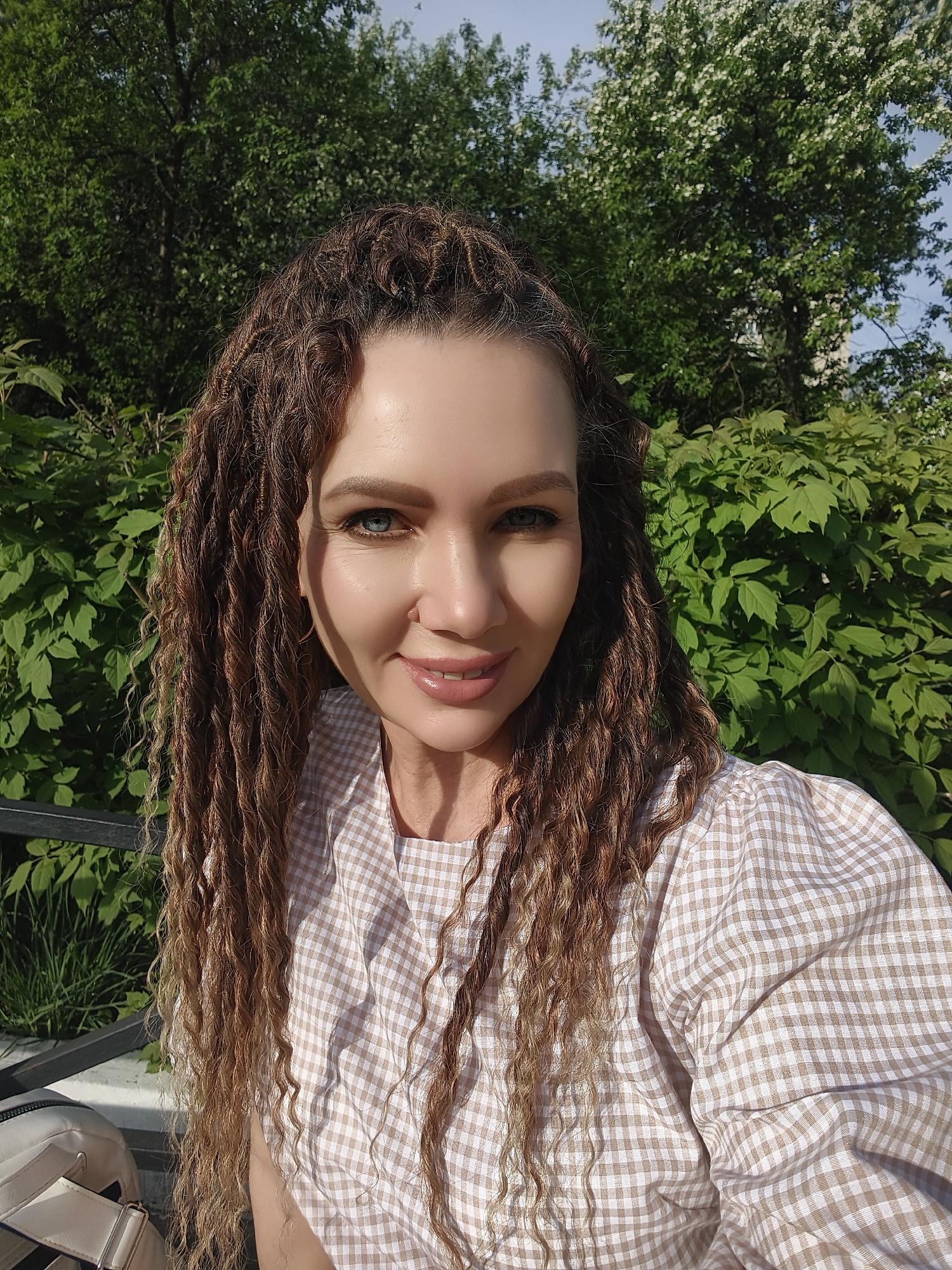 A woman is captured in a medium shot from the chest up, appearing to be outdoors in the city of Unknown, Unknown. She has long, brown, textured hair falling around her face and shoulders. She wears a light-colored, checkered shirt and has light blue eyes. She is smiling. Behind her is a backdrop of green foliage from bushes and trees. The lighting suggests it is daytime, with some areas of her face in shadow. A portion of a dark-colored bench is visible on the left side, along with a light-colored bag.