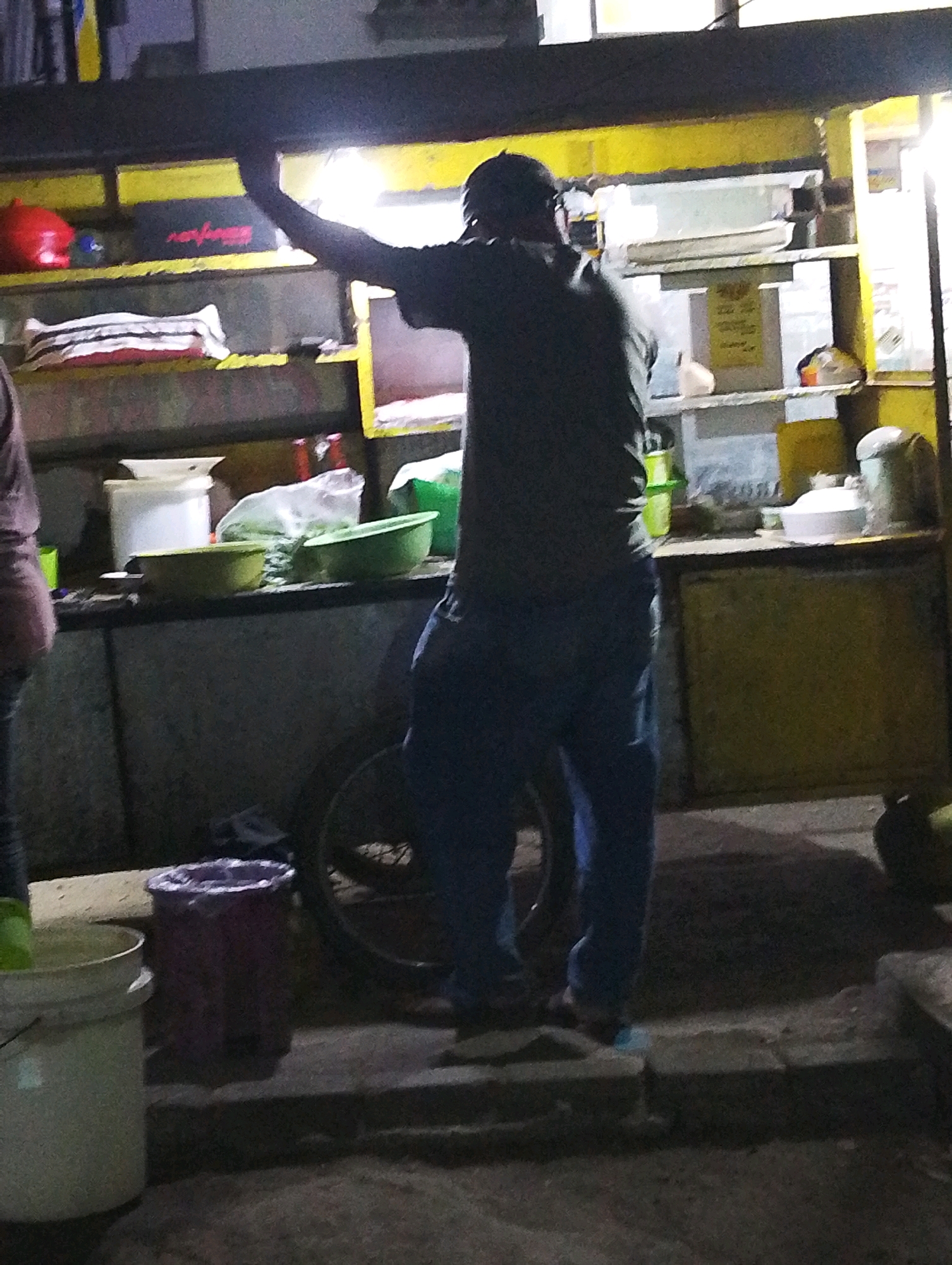 A person is standing in front of a food stall at night in Manaruwi, Indonesia. The person is wearing a dark shirt and blue pants. They are standing next to a bicycle wheel. The food stall has yellow shelves and a dark counter. There are various items on the shelves, including bowls, containers, and what appears to be food. There are also several buckets in front of the stall. The scene is dimly lit by artificial lighting.