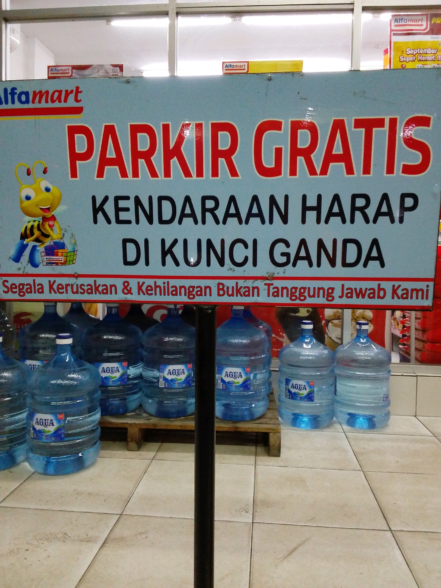 The image depicts a sign in front of several large bottles of Aqua brand water, likely in a retail setting. The sign, in Indonesian, reads "PARKIR GRATIS KENDARAAN HARAP DI KUNCI GANDA" which translates to "FREE PARKING VEHICLES PLEASE DOUBLE LOCK". Below this, it states "Segala Kerusakan & Kehilangan Bukan Tanggung Jawab Kami" meaning "All Damage & Loss Is Not Our Responsibility". The sign features a cartoon bee mascot associated with Alfamart, a convenience store chain in Indonesia. The Aqua bottles are typical large refillable containers. The flooring appears to be tiled. While the image was taken in Deli Tua, Indonesia, specific details about the environment beyond the immediate foreground and sign are not clearly discernible. There are no people or direct interactions visible in the image. The scene suggests a parking area or entrance to a store where customers are being reminded to secure their vehicles.