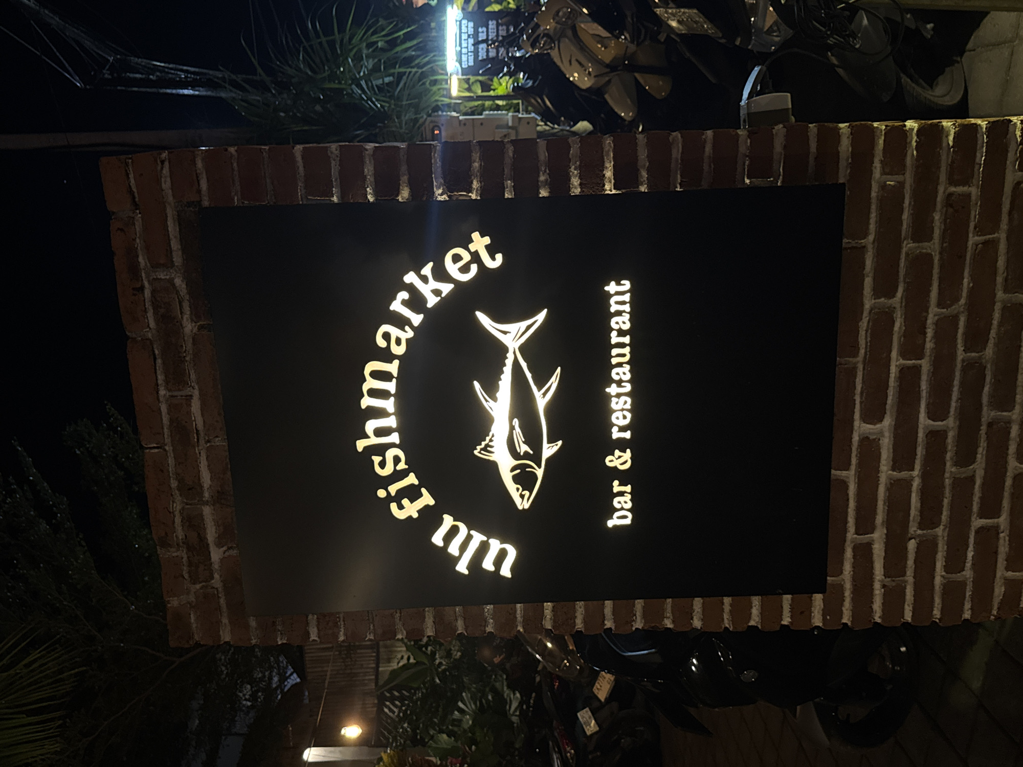 A sign for "Ulu Fishmarket, bar & restaurant" in Kuta Selatan, Indonesia, is displayed on a brick wall. The sign is black with gold lettering and a stylized fish graphic. There are motorcycles parked around the base of the sign. The photograph was taken at night.