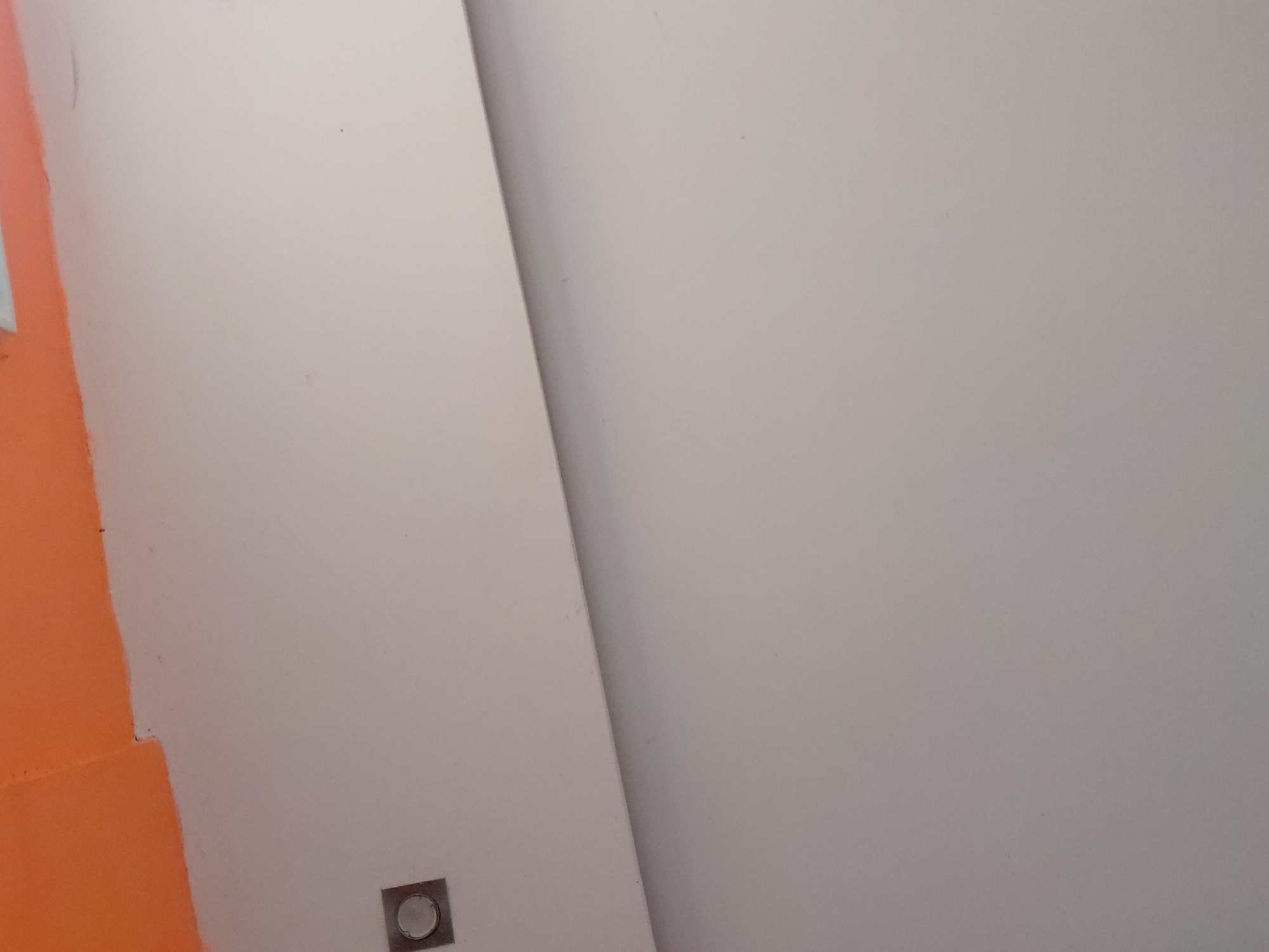 The image presents a close-up, abstract view of an interior wall or architectural surface. The dominant color is a light, off-white or very pale grey, covering most of the frame. On the left side, a section of the surface is painted a vibrant, bright orange, creating a sharp vertical and angled color division. The boundary between the orange and light grey paint is generally straight but exhibits minor irregularities where the colors meet.
Near the bottom-left corner, within the light grey area and adjacent to the orange, a small, square, metallic-colored object is visible. This object features a circular, possibly translucent or reflective, center, suggesting it could be a flush-mounted light fixture, a sensor, or a button.
A subtle diagonal line or shadow runs from the upper-middle section of the image down towards the bottom-right, indicating a possible corner, a fold in the surface, or a shift in plane. The lighting appears even, without harsh shadows, suggesting ambient or indirect light. No people or activities are depicted. There is no visible text. The scene is static, focusing on color, texture, and geometric divisions within an architectural context in Santa Ana, Venezuela.