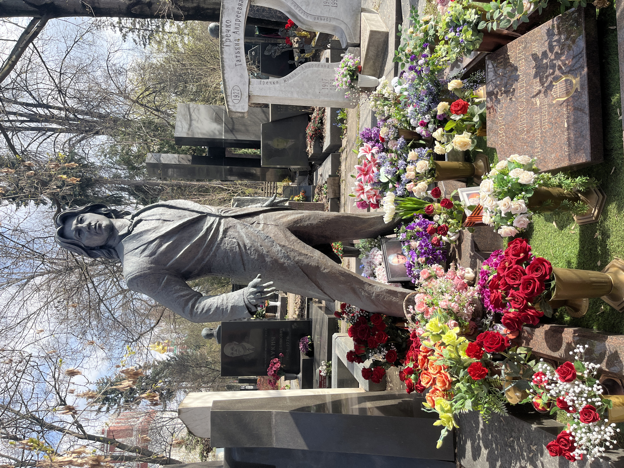 The image shows the grave site of Dmitry Hvorostovsky, the Russian opera singer, at the Novodevichy Cemetery in Moscow, Russia. The grave is marked by a life-size statue of Hvorostovsky in a suit and is covered with flowers and wreaths. A portrait of an older woman is visible among the flowers, as well as several tombstones with inscriptions including "Лукич" (Lukich), "Гречко" (Grechko), and "ХВОРОСТОЛСКИ" (Hvorostovsky). The surrounding environment appears to be a cemetery with many other graves, trees, and foliage.