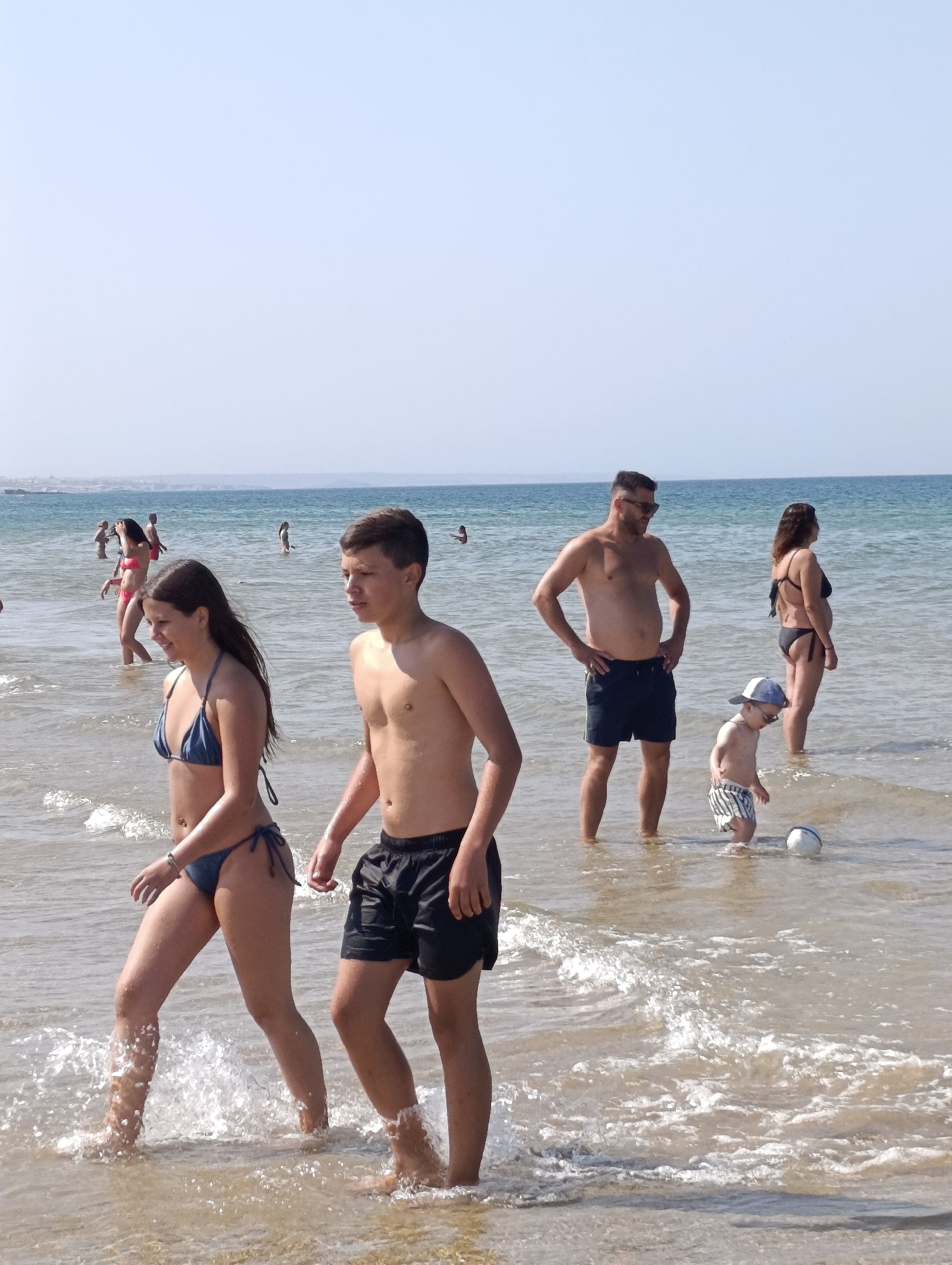 This is a photograph taken at a beach in Sines, Portugal. The scene depicts several people enjoying the ocean on a sunny day, likely in the afternoon, given the bright light and clear sky. The water is relatively calm with small waves breaking on the shore.
In the foreground, a young woman and a teenage boy are walking through the shallow water. The woman is wearing a blue bikini, and the boy is wearing black swim shorts. Both appear to be casually enjoying their time at the beach.
Further in the background, a man in dark blue swim shorts and sunglasses stands with his hands on his hips, looking towards the water. Beside him, a young child in striped swim shorts and a white cap is playing with a white ball in the water. A woman in a black bikini stands to the right of the man.
Other people can be seen in the water and on the shore in the distance, suggesting a moderately busy beach day. The overall atmosphere seems relaxed and summery. The horizon shows a faint line of land in the distance, typical of a coastal view. There is no visible text in the image.