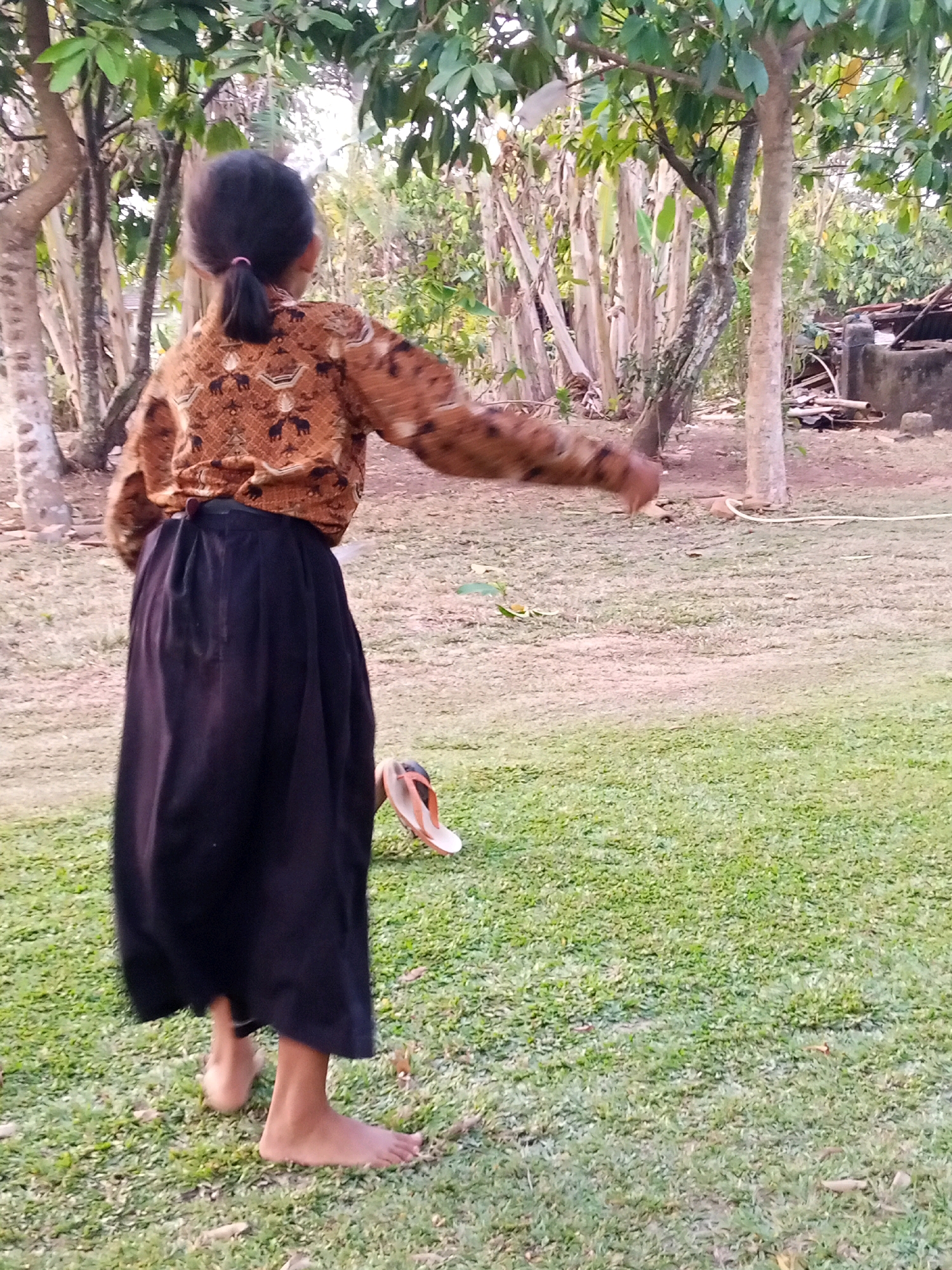 In an outdoor, rural setting in Unknown, Indonesia, a young person stands with their back to the camera, facing away from the viewer. The individual is barefoot and wears a long, dark skirt and a patterned, long-sleeved shirt in shades of brown and tan. The shirt features a repeating design, possibly of elephants. The person's dark hair is pulled back into a ponytail with a pink tie. They are in motion, with their right arm extended outwards, suggesting they are in the process of performing an action, possibly throwing something or playing a game. Near the person's feet, on the grassy ground, lies a pair of orange and white flip-flops. The environment is natural, with several trees and foliage visible in the background, creating a sense of a village or wooded area. The ground is covered in green grass, with some fallen leaves scattered about. The lighting suggests it is daytime, possibly late afternoon, with soft, diffused light.