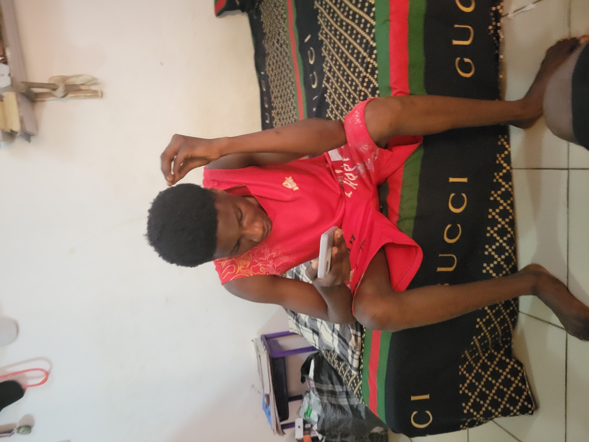 A young Black male is sitting on a bed and looking at his phone. He is wearing a red sleeveless shirt with a yellow design on it and red shorts. The bed is covered with a black sheet that has a gold pattern and the word "GUCCI" printed on it in gold letters. There is also a red and green stripe on the bed. In the background, there is a white wall and a small desk with some items on it. The room appears to be dimly lit.