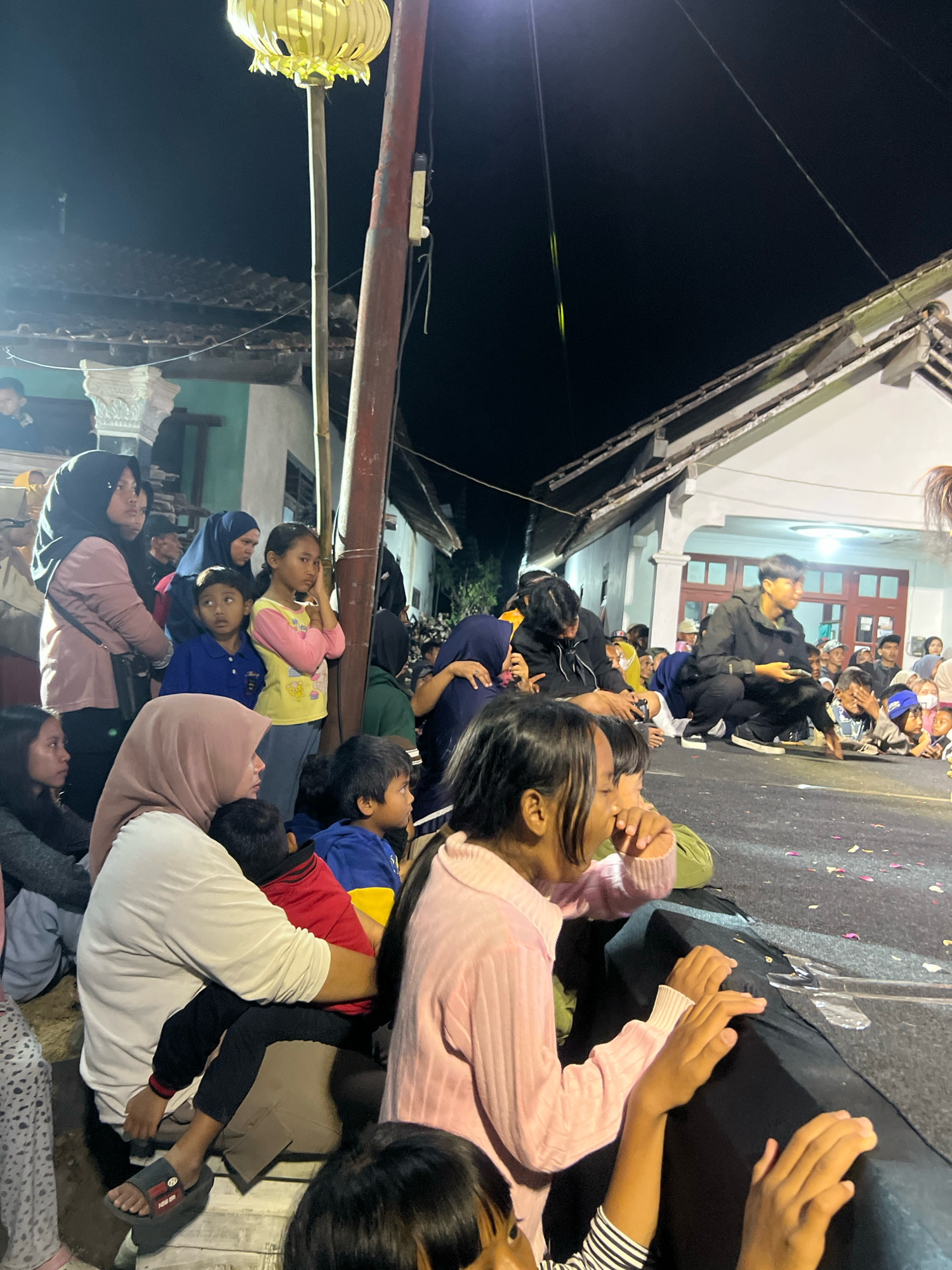 In the city of Madyocondro, Indonesia, an outdoor nighttime gathering is in progress. The scene is illuminated by a large, hanging yellow lantern affixed to a pole, casting a warm glow on the surrounding area. A crowd of people, including adults and children, are present, many of whom are looking towards an unseen focal point, suggesting an ongoing event or performance. Several individuals are seated or squatting on the ground, while others stand. Buildings with tiled roofs are visible in the background. The ground is paved, with some debris, possibly flower petals, scattered across it. The overall atmosphere appears to be one of communal engagement and observation in a local setting.
