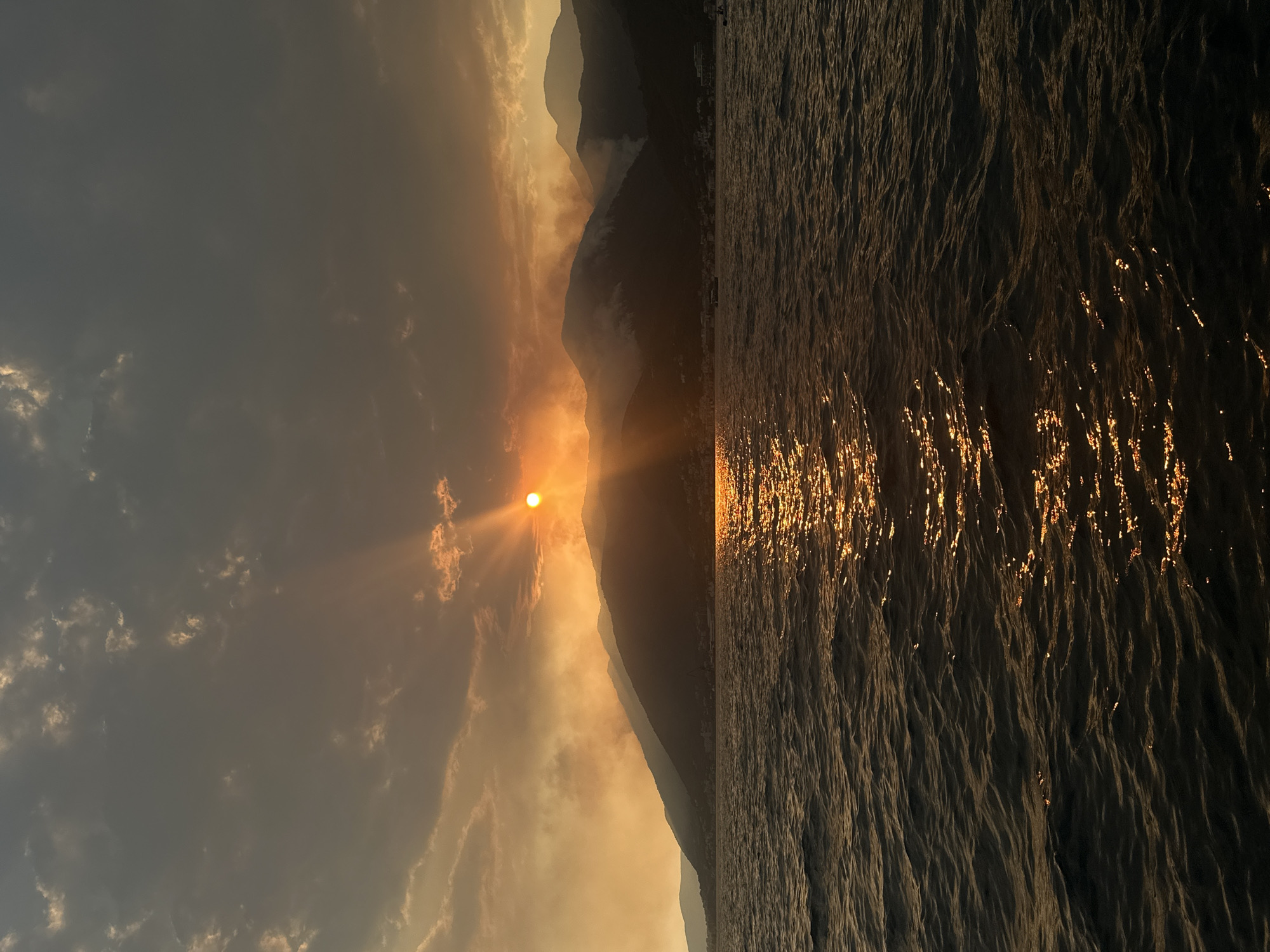 The image depicts a sunset over the Bay of Kotor, as seen from the water near Donja Lastva, Montenegro. The sun, a brilliant orange orb, is partially obscured by clouds and hovers just above a silhouette of mountainous terrain. Rays of light pierce through the clouds, creating a dramatic atmospheric effect. Smoke or haze rises from the slopes of the mountains, possibly indicating a fire or industrial activity. The foreground is dominated by dark, choppy water, reflecting the warm hues of the sunset. Along the base of the mountains, faint lights suggest a coastal settlement. The overall scene is one of natural beauty, with a touch of environmental concern due to the visible smoke.