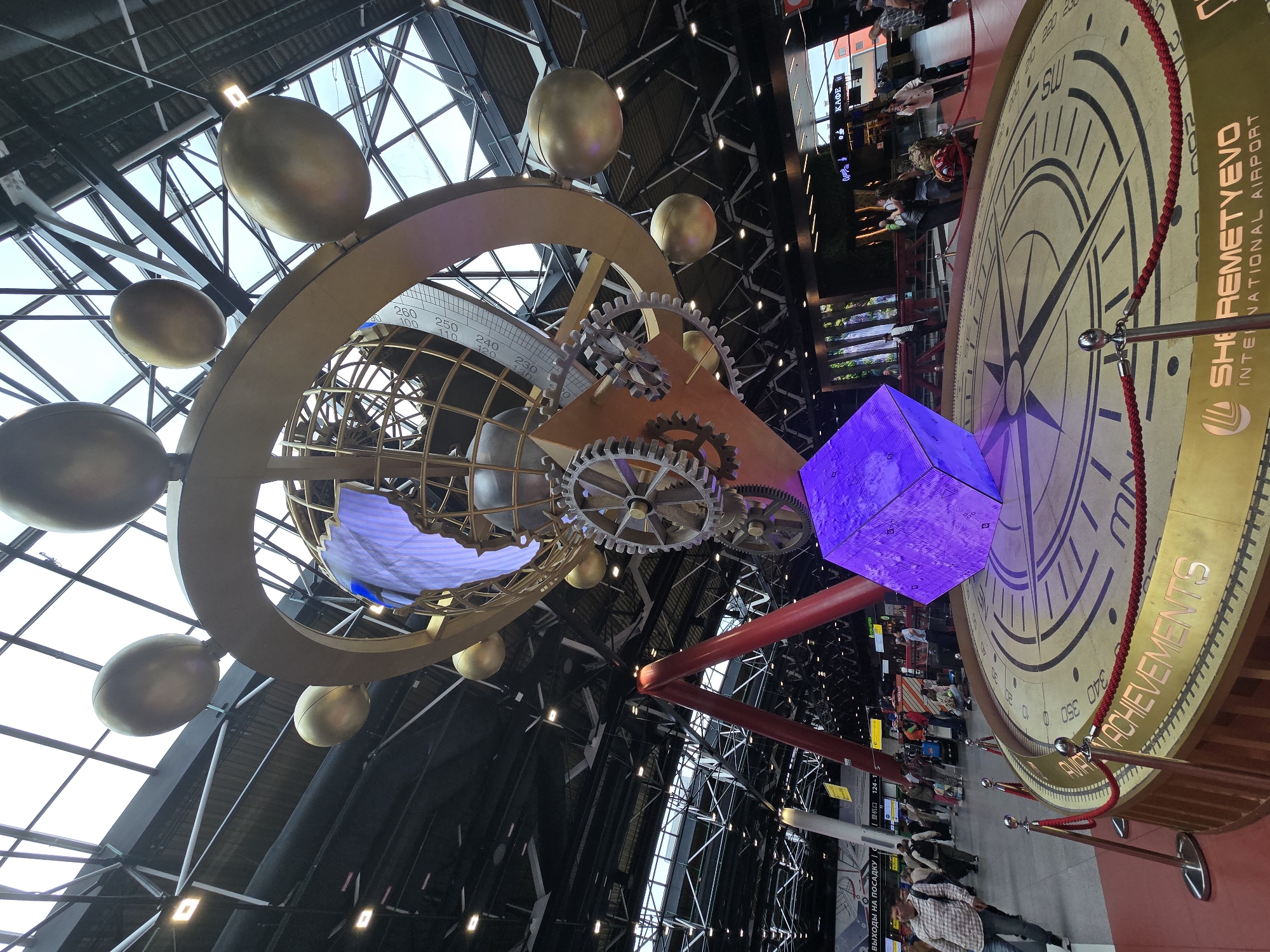 This image captures the interior of a brightly lit, modern airport terminal, identified by visible text as Sheremetyevo International Airport in Khimki, Russia. The setting is characterized by a high, geometric glass and metal ceiling structure, allowing ample natural light to illuminate the space, suggesting it is daytime.
The central focus is a monumental, multi-component art installation. Suspended from the ceiling is a large, golden-hued structure resembling an armillary sphere or a complex celestial globe, complete with intricate rings, visible gears, and several large, golden, spherical or ovoid elements that evoke planets or moons. Below this, a massive circular display is set into the floor, functioning as a large compass rose or clock face with directional markings like "SW" and numerical degrees (e.g., 260, 350, 0, 100). This floor display is cordoned off by red velvet ropes and stanchions. A vibrant, glowing purple cube, possibly a digital display or light fixture, stands near the compass rose, connected by a prominent, angular red structural beam.
Numerous people are visible throughout the terminal, primarily in the background and around the floor display. They appear to be travelers, walking, standing, or waiting, consistent with the bustling environment of an international airport. No strong emotions are discernible on their faces; their attire is casual.
Notable textual cues confirm the location and context: "SHEREMETYEVO INTERNATIONAL AIRPORT" and "ACHIEVEMENTS" are prominently displayed on the large circular floor installation. Other signs include "КАФЕ" (Cafe) and "ВЫХОДЫ НА ПОСАДКУ" (Boarding Gates) in Cyrillic, along with Chinese characters "登机口 124-1" (Boarding Gate 124-1), indicating its international status.