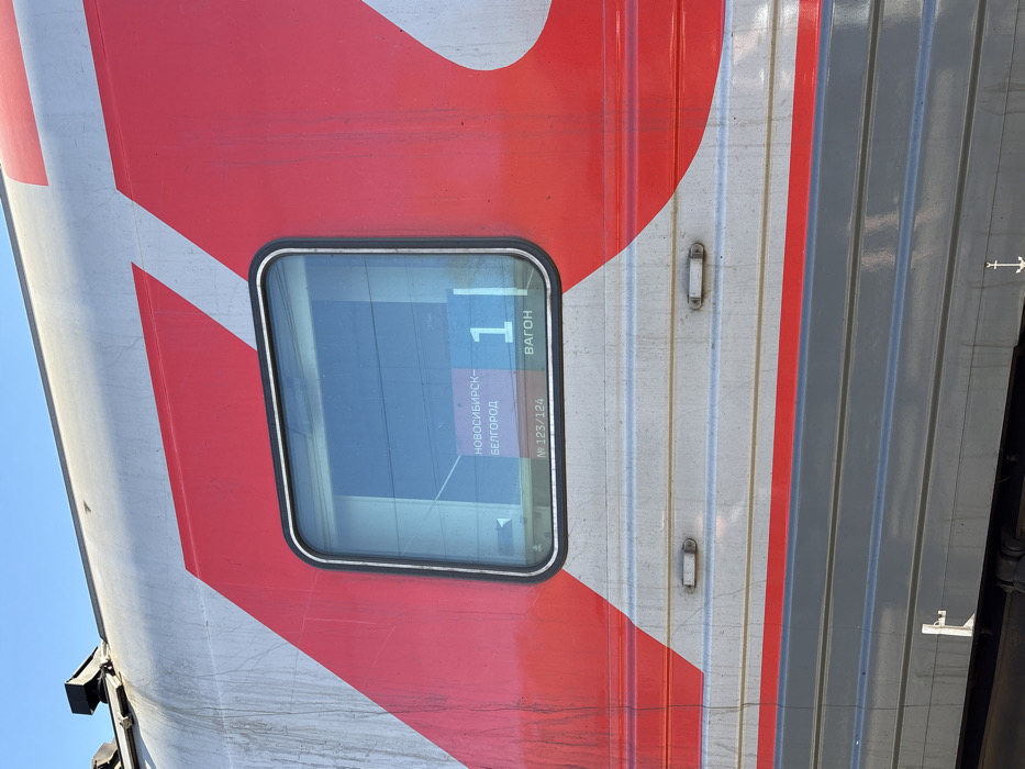 The image presents a forensic view of the exterior side of a passenger train car, stopped in Kropachyovo, Russia, under a clear blue sky. The upper section of the train car is silver-grey, adorned with large, stylized red branding, likely associated with Russian Railways (RZD). A dark grey lower section is separated by a thin red horizontal stripe. A rectangular window with rounded corners is visible, featuring an internal display. This display shows route information in Russian: "НОВОСИБИРСК-БЕЛГОРОД" (NOVOSIBIRSK-BELGOROD) as the destination, "1" as the wagon number, and "№ 123/124" as the train number, along with the word "ВАГОН" (VAGON). The train's exterior exhibits visible streaks of dirt and weathering, particularly on the silver-grey panels. Below the window, two small, rectangular light fixtures are embedded in the side. Technical markings are present on the dark grey lower panel. No individuals or discernible actions are observed in the scene.