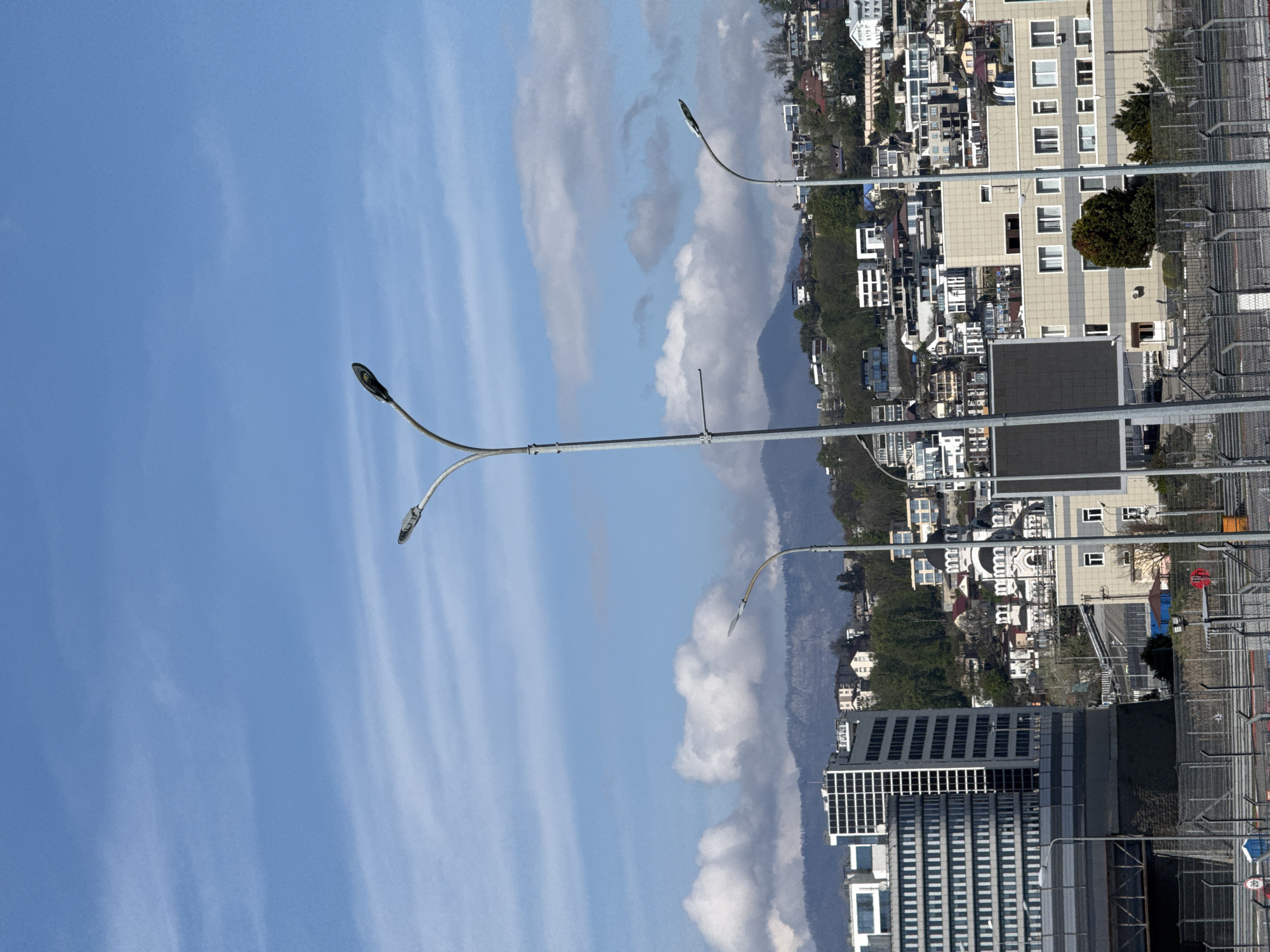 The image displays a sunlit urban landscape with a distinct mountainous backdrop, viewed under a clear blue sky. The perspective is from a slightly elevated vantage point, showcasing a sprawling city with diverse architecture.
Two prominent, modern street light poles with curved arms, each holding two light fixtures, dominate the upper and middle sections of the image, one positioned on the left and the other on the right. The sky above is a bright, clear blue, adorned with streaky white cirrus clouds and some larger, fluffy cumulus clouds gathered closer to the horizon.
A range of substantial mountains or hills forms a natural boundary across the mid-ground, separating the expansive sky from the dense urban development. These mountains exhibit a mix of green vegetation and rocky outcrops.
The city occupies the lower half of the frame, extending from the base of the mountains towards the viewer. On the right side, a large, multi-story building with a light-colored facade and numerous rectangular windows is visible, partially obscured by a metal fence structure in the immediate foreground. To the left, a tall, modern building with a distinctive grid pattern of dark windows is prominent in the lower-left corner. Beyond these structures, a multitude of buildings, including both detached houses and larger apartment blocks of varying architectural styles, populate the urban area, extending up the slopes of the hills. Green trees and foliage are scattered throughout the built environment.
No people are visible in the scene. The time of day is clearly daytime, indicated by the bright illumination and the blue sky, suggesting fair weather conditions. Location cues include the combination of dense urban development, multi-story buildings, and proximity to mountains. Visible text includes a blue rectangular speed limit sign displaying "40" in white within a red circle, and a red circular sign with a diagonal white line, both partially seen on the right side near the fence.