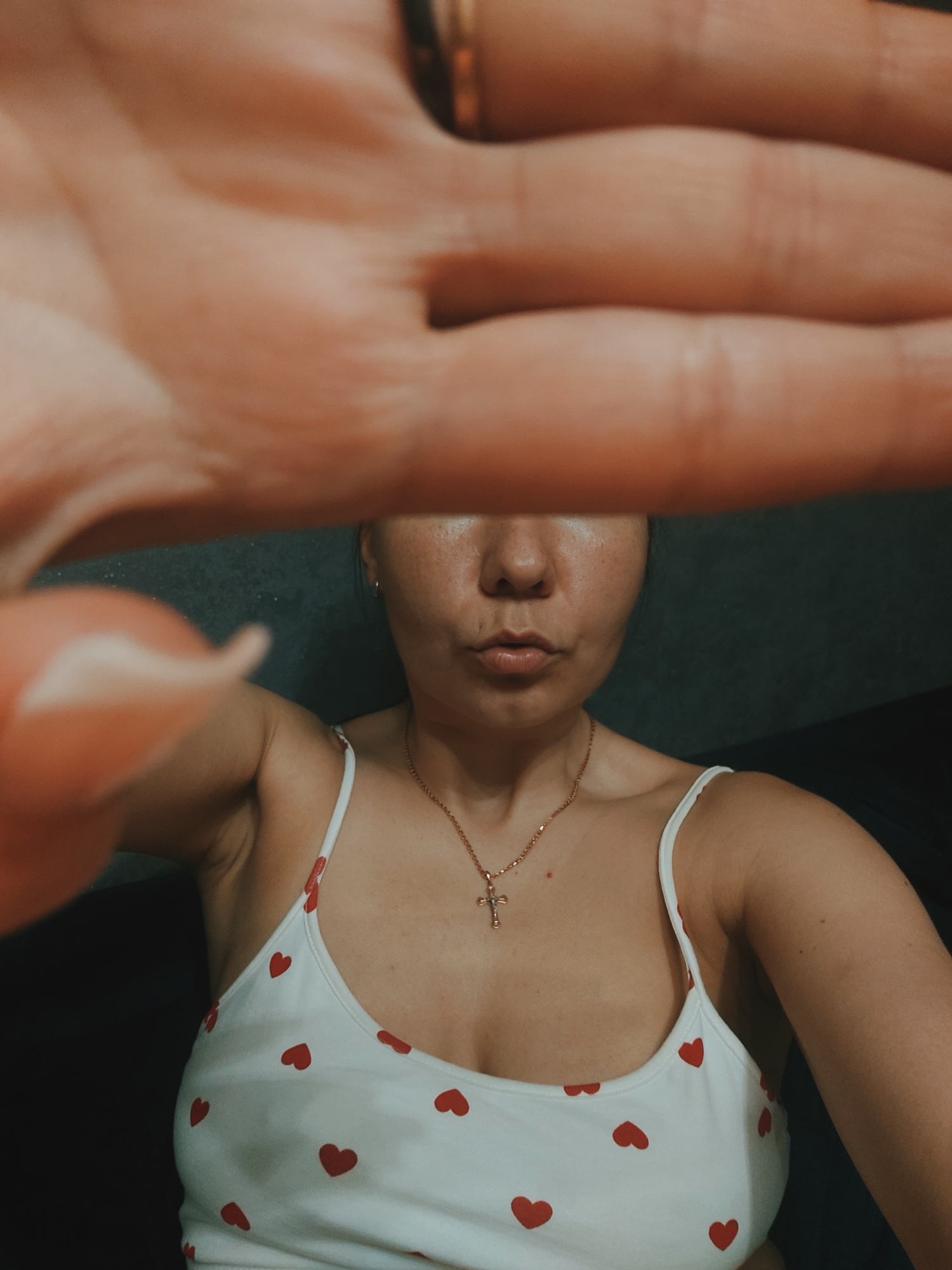 A young woman is captured in a close-up selfie, with her right hand extended and blurred across the top of the frame, obscuring a portion of her face. She is wearing a white tank top adorned with small red hearts and a gold cross necklace. Her expression is suggestive, with her lips pursed. The background is dark and out of focus, with hints of a textured surface. The lighting is dim, creating a moody atmosphere. No specific location in Chaikovsky, Russia, can be determined from the visual information provided.