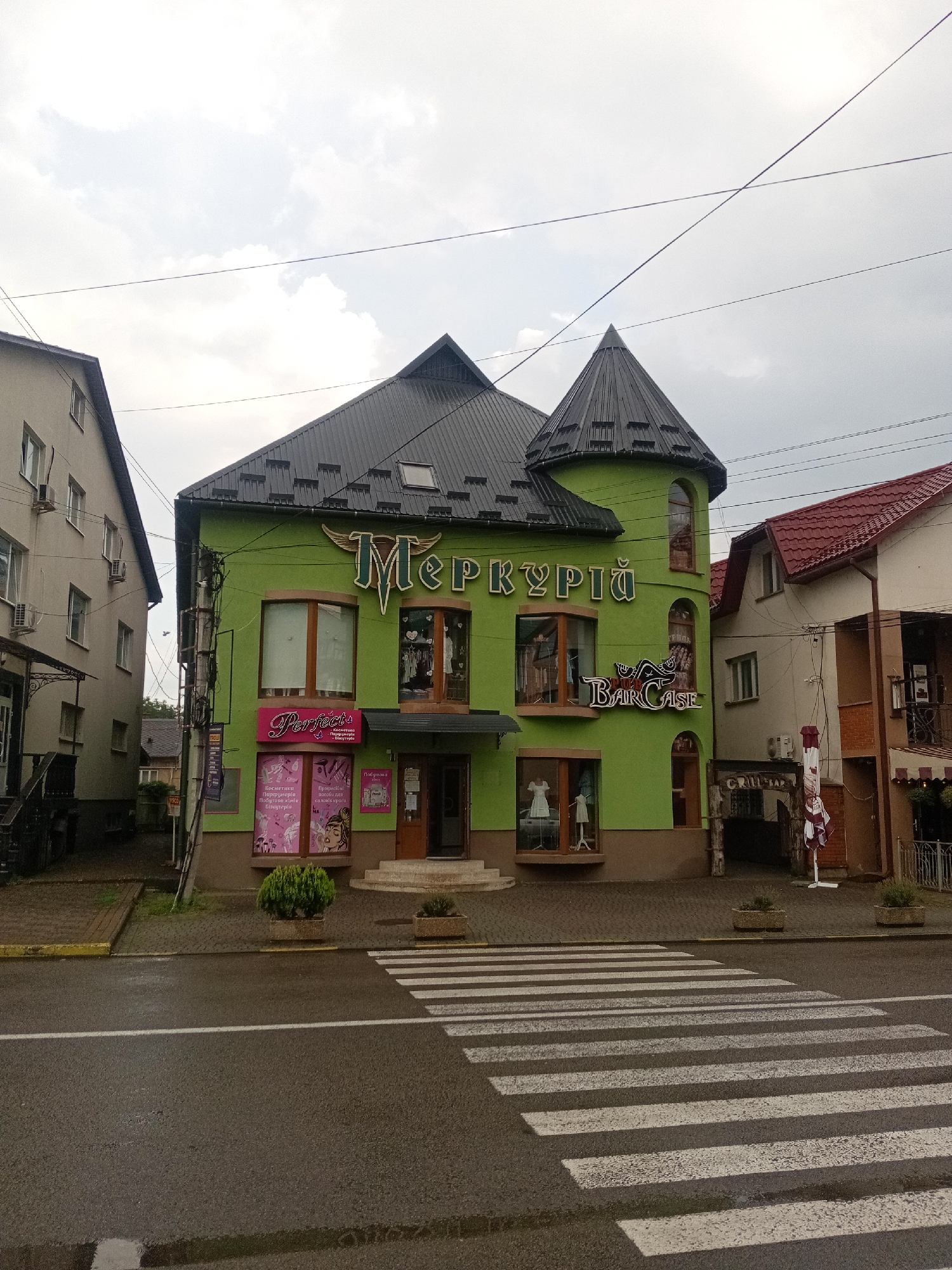 The image shows a two-story building with a bright green facade in Tiachiv, Ukraine. The building has a distinctive dark gray, multi-pitched roof with a conical turret on the right side. The facade features several windows, some with wooden frames, displaying mannequins and clothing. A sign above the main entrance reads "МЕРКУРІЙ" in large, stylized letters. Below this, a smaller sign says "BAR CASE". The building is situated on a street with a pedestrian crossing in the foreground. The sky is overcast. To the left of the green building is a lighter-colored, multi-story building, and to the right, another building with a red tiled roof. The scene appears to be a commercial area on a street.