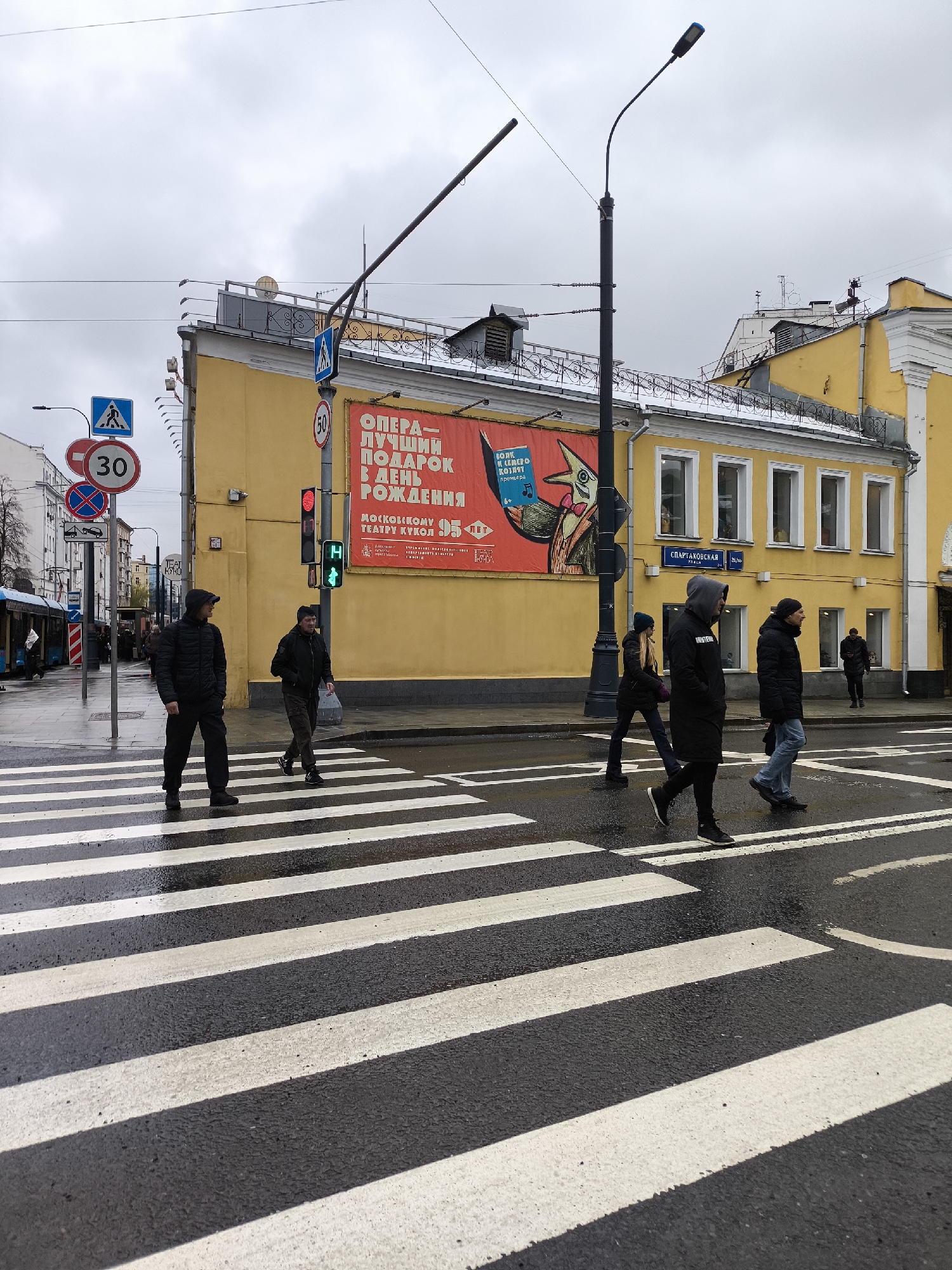 The image captures an urban street scene in Moscow, Russia, on an overcast day. Several pedestrians are depicted using a zebra crossing on a wet asphalt road, suggesting recent rain. The traffic light for pedestrians is green, allowing them to cross.
On the left side of the crosswalk, a man in a black puffer jacket and dark pants walks towards the right. Another man, wearing a dark hoodie and jacket with blue jeans, crosses further right. A woman in a dark jacket and beanie is walking from left to right, behind the man in the dark hoodie. Further in the background, another man in a dark jacket is visible on the sidewalk.
The background features a prominent yellow building with multiple windows on its upper floor. A large red banner covers a significant portion of its facade, displaying text in Russian. Above the building, the sky is grey and cloudy. Various street furniture is present, including street lamps, utility poles with wires, and several street signs. A bus stop or trolleybus stop structure is visible on the far left.
Visible text includes multiple street signs: a speed limit of "30," a partially obscured "50," and several pedestrian crossing signs. The red banner on the building reads "ОПЕРА - ЛУЧШИЙ ПОДАРОК В ДЕНЬ РОЖДЕНИЯ" (Opera - The Best Birthday Gift), "МОСКОВСКОМУ ТЕАТРУ КУКОЛ" (Moscow Puppet Theatre), and "95," likely indicating an anniversary. Another section of the banner advertises "ВОЛК И СЕМЕРО КОЗЛЯТ премьера" (The Wolf and the Seven Kids premiere). A blue street sign on the yellow building reads "СПАРТАКОВСКАЯ 26/10" (Spartakovskaya 26/10), identifying the street. Another small sign near the bus stop says "TEATR KUKOL". The wet road surface and grey sky are notable details.