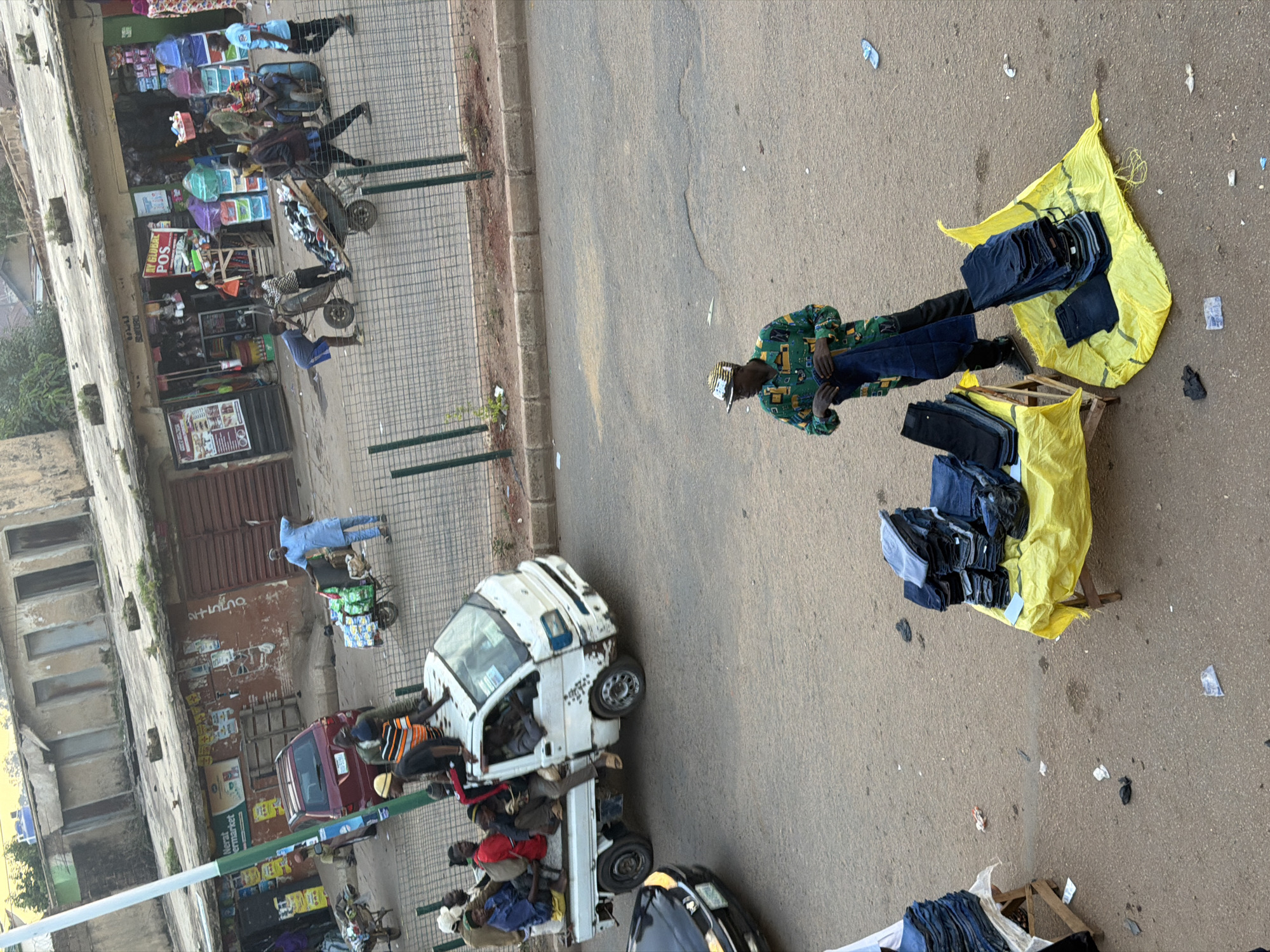 A street scene in Bukuru, Nigeria, shows an outdoor market with several people and stalls. In the foreground, a man wearing a patterned green and yellow shirt and a striped hat is displaying folded jeans. He is standing behind a yellow tarp on which more jeans are neatly stacked. To the left, another stall with a wooden frame displays stacks of folded jeans on a white sheet.
Further in the background, a white pickup truck is loaded with people. The truck is on a paved street, and a red SUV is parked nearby. Buildings with weathered facades line the street, some with open storefronts. A green metal fence separates the market area from the street. A person in a blue tunic is seen pushing a cart loaded with packaged goods. The overall scene depicts a busy, informal commercial environment.